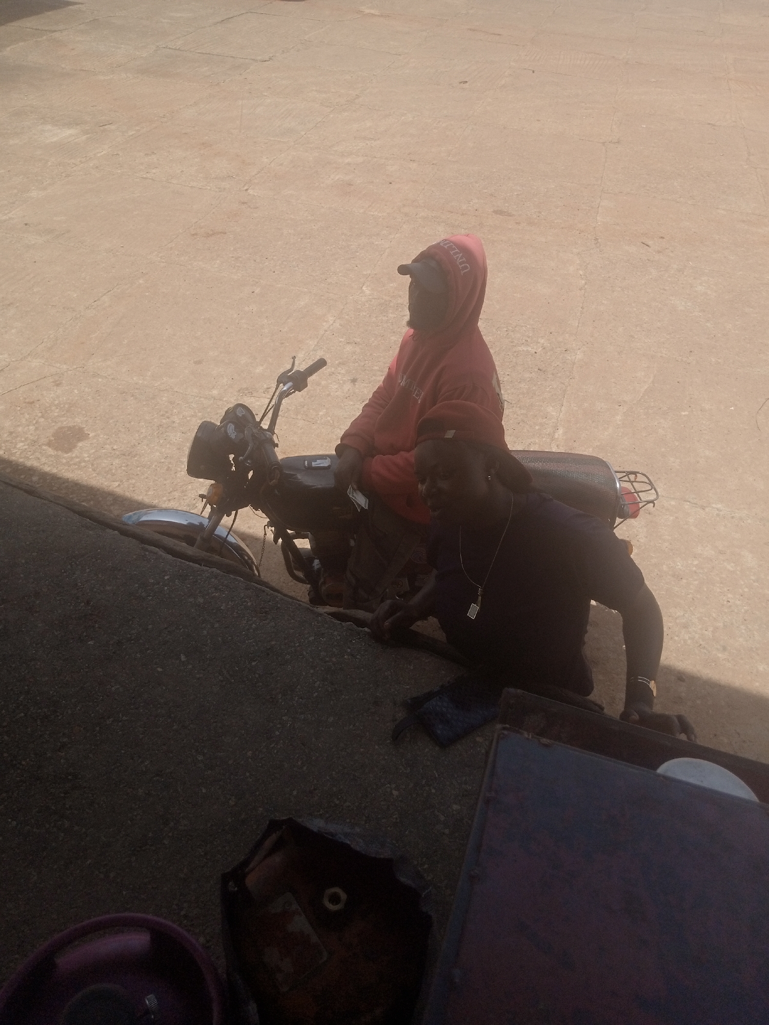 Two people are seen in the image, both wearing casual clothing. One person is wearing a red hooded sweatshirt and a dark baseball cap, while the other is wearing a dark t-shirt, a dark baseball cap, and a necklace. They are both on or near a motorcycle. The motorcycle is dark in color with a chrome fender and handlebars. The scene appears to be outdoors, with a paved surface that looks like concrete or asphalt. The lighting suggests it is daytime, possibly mid-morning or afternoon, with shadows cast on the ground. The overall impression is a casual outdoor setting. There is some text visible on the red hoodie, though it is partially obscured and difficult to read clearly.
