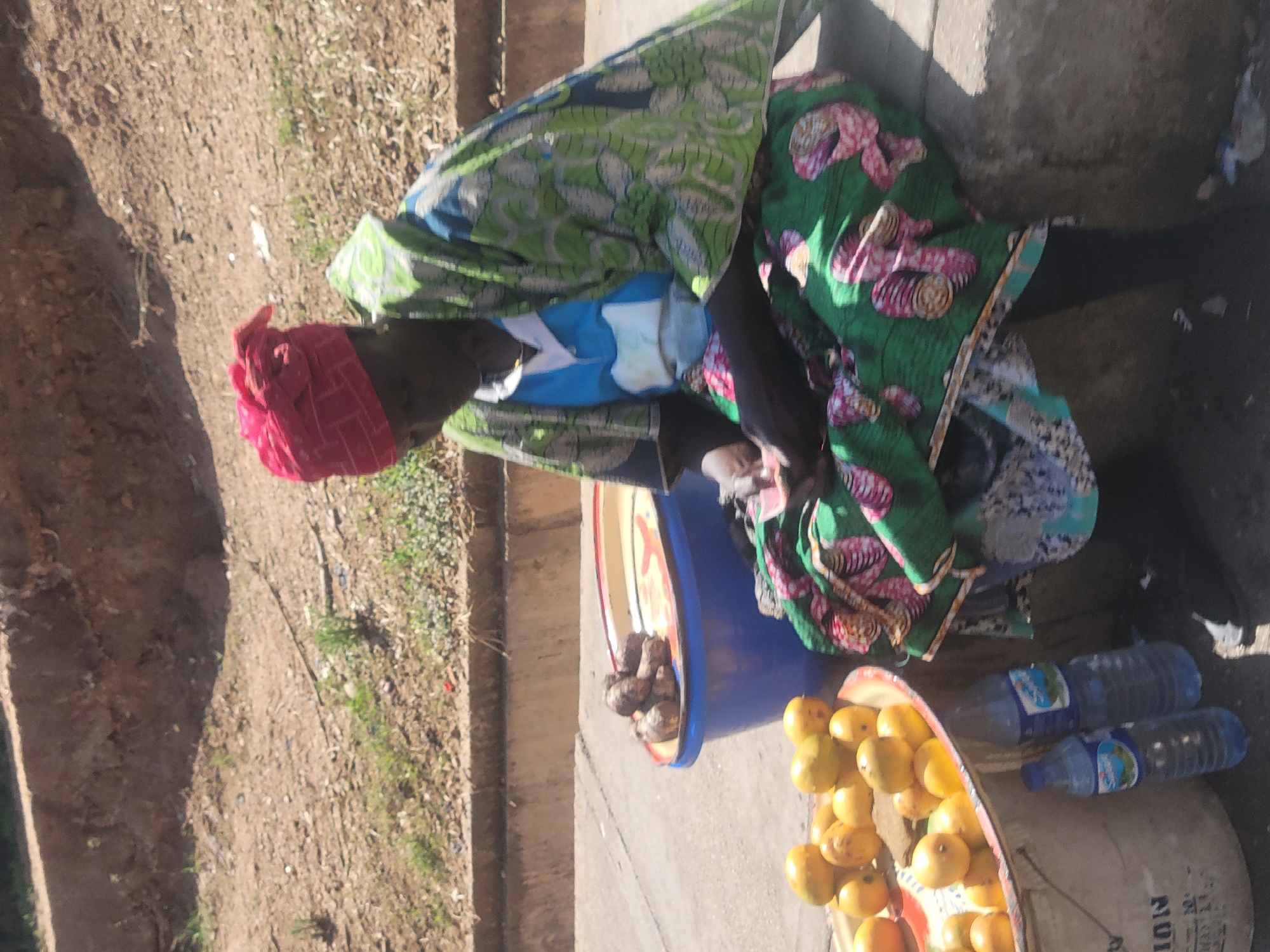 A woman wearing a red headscarf, a blue shirt, and a green patterned wrap sits on the pavement in Jos, Nigeria. She has two trays in front of her: one with yellow fruit, the other with brown produce. There are two bottles of water on a pail next to the fruit. The area next to the pavement is bare soil.