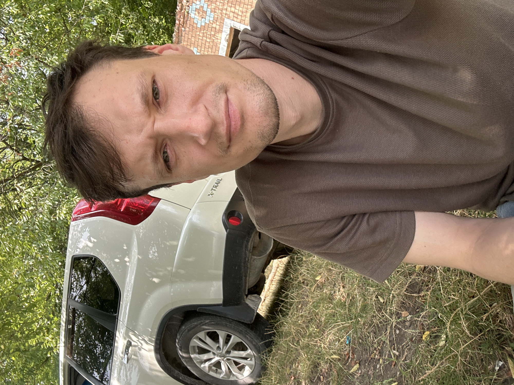 A man is taking a selfie in front of a white Nissan X-Trail SUV parked on a grassy area with trees in the background. The man is wearing a brown t-shirt. A brick wall is visible behind him. The location appears to be Makiv, Ukraine, based on the context provided.
