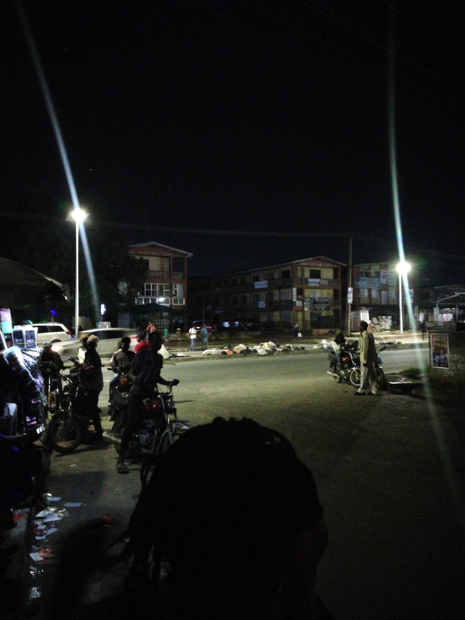 The image shows a street scene at night in Lagos, Nigeria. Several people are present, some on motorcycles. There are buildings in the background illuminated by streetlights. Trash is scattered along the side of the road. One person is standing on the side of the road. The scene has a sense of urban activity and possibly commerce.