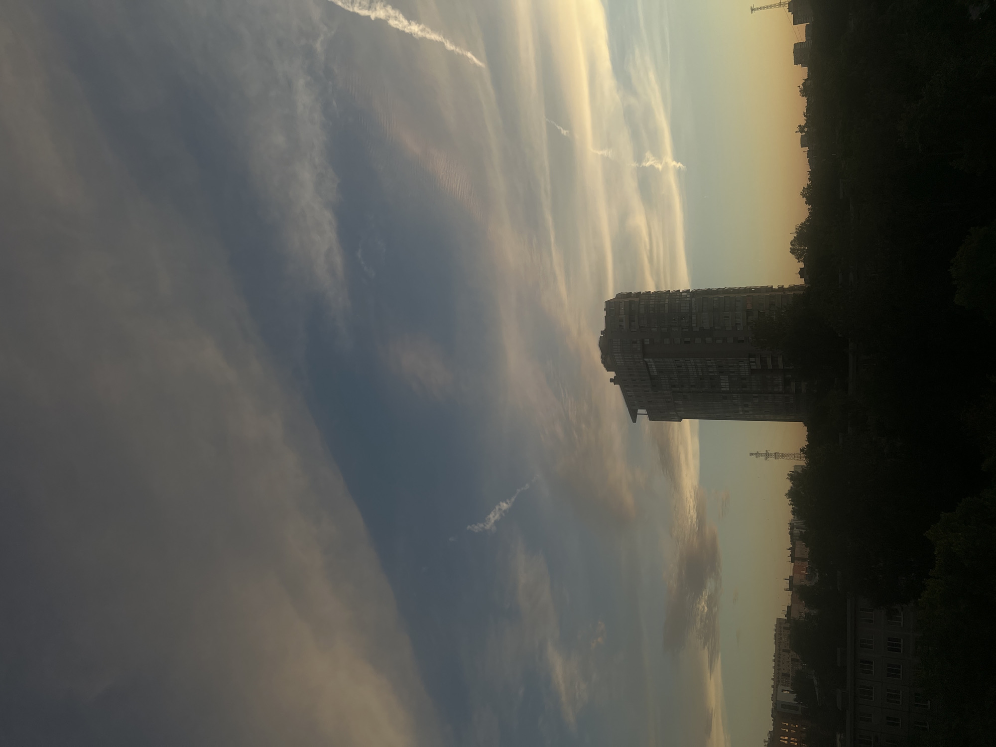 An urban cityscape is depicted under a dramatic sky filled with wispy, elongated clouds, characteristic of sunset or sunrise. A tall, modern apartment building dominates the center-right of the frame, its rectangular structure and numerous windows clearly visible. In the foreground and middle ground, silhouetted trees and lower-rise buildings form a dark, dense layer. The background reveals a distant skyline with several more buildings, and a telecommunications tower stands prominently. The sky transitions from a pale yellow and orange near the horizon to a deep blue overhead, with faint contrails visible. The scene appears to be a view from an elevated position, looking out over a residential area. Petro-Slavyanka, Russia, is the specified location. There are no people or animals visible in the image.
