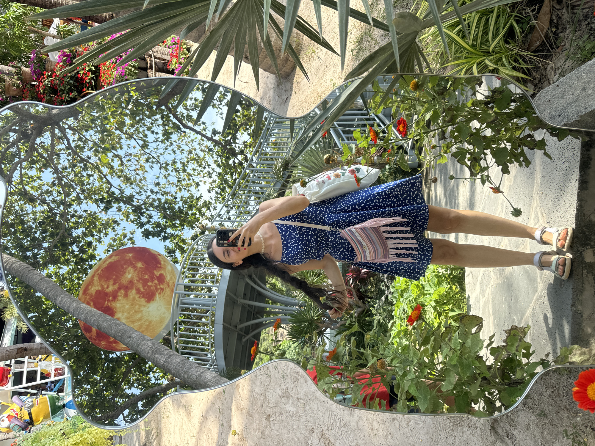A woman is taking a selfie in a wavy, guitar-shaped mirror located in a garden in Mũi Né, Vietnam. The woman is wearing a blue dress with white polka dots, a pearl necklace, and white sandals. She is holding her phone in her right hand and her long dark hair in her left hand. She has a fringed purse slung over her left shoulder and a white tote bag with floral print hanging on her right. In the mirror's reflection, a large, red, textured sphere resembling the moon is visible behind a spiral staircase. Lush green trees with sunlight filtering through their leaves and a bright blue sky are also reflected in the mirror. The garden is filled with various plants, including flowering bushes with red and pink blooms, palm-like plants, and green foliage. The ground is paved with stone.