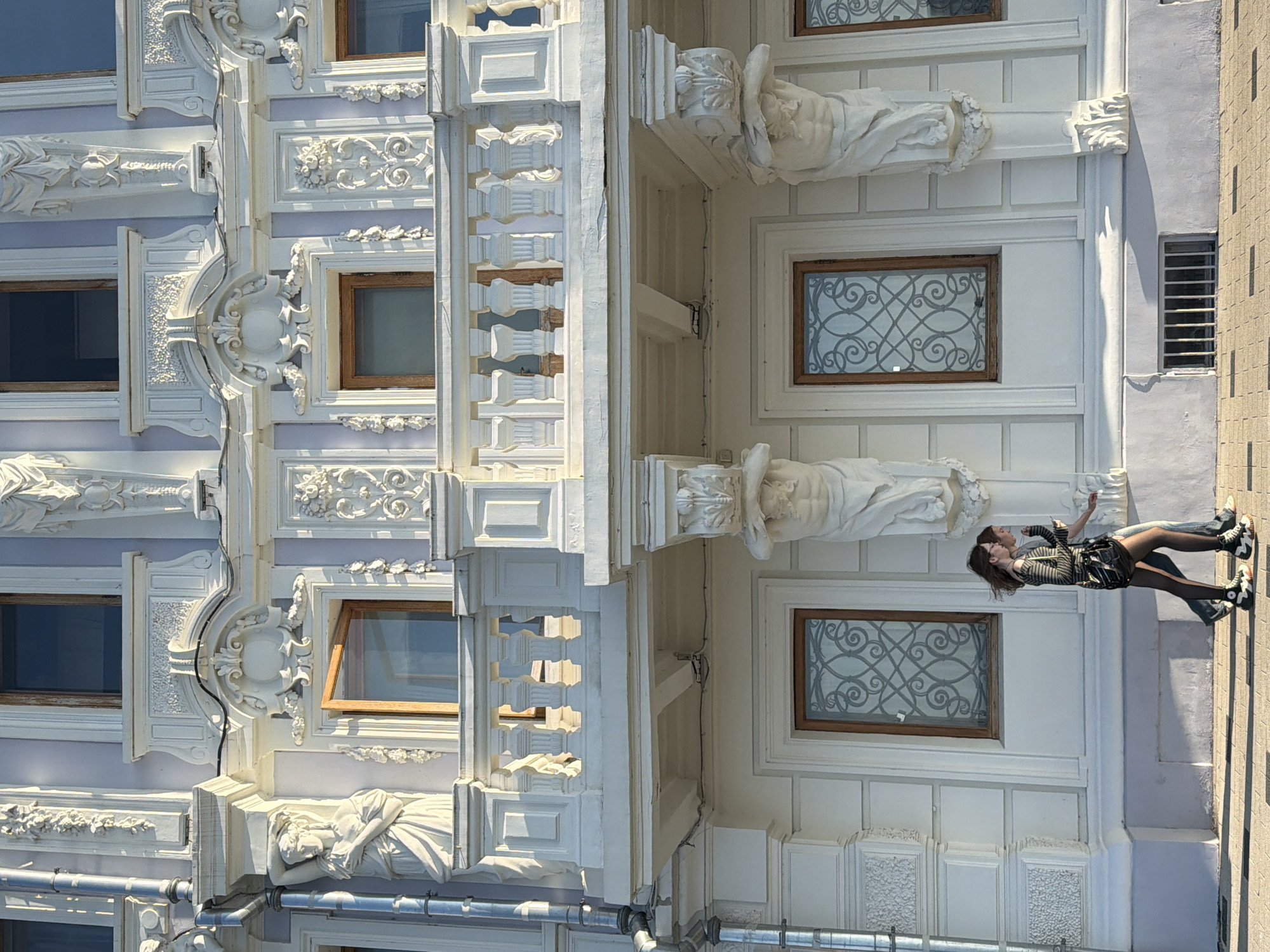 The image captures a street scene in Nizhny Novgorod, Russia, focusing on the ornate facade of a building. The building features intricate white stucco detailing, including sculptures of Atlas figures supporting balconies and decorative reliefs. Multiple windows are visible, some with wooden frames and decorative grilles. Two young women are walking along the sidewalk in front of the building, one holding a phone and the other carrying a bag. The sun casts shadows on the facade and the ground, indicating a bright day. A metal drainpipe runs down the side of the building.