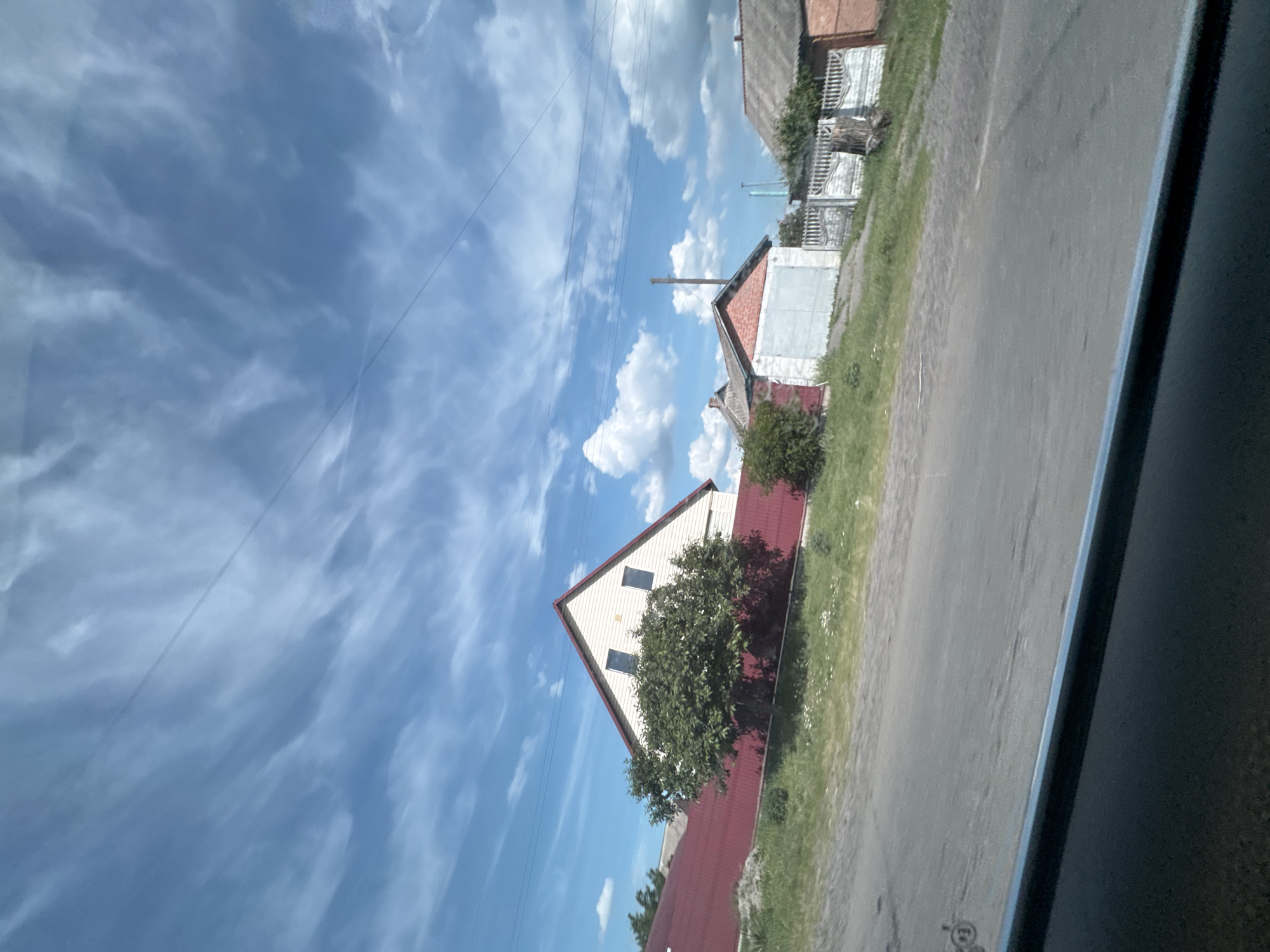 The image captures a rural or semi-rural residential street scene in Markivka, Ukraine, viewed from inside a vehicle, indicated by the dark car interior visible at the bottom. The sky, occupying the upper two-thirds of the frame, is clear blue with scattered white clouds, ranging from wispy cirrus-like streaks in the upper left to more defined cumulus clouds toward the right. Multiple thin power lines traverse the sky horizontally.
The foreground features a grey asphalt road, partially visible, with a grassy verge bordering the properties. No people are present.
On the left side of the midground, a two-story house with light-colored siding and a steep red gable roof is prominent. Two dark, rectangular windows are visible on its upper facade. A dense green deciduous tree stands directly in front of this house, partially obscuring its lower section. Behind the tree and house, a long red fence or wall extends horizontally.
To the right, further along the road, a brick outbuilding with a grey garage door and a dark roof is visible. A white, ornate fence with decorative balusters runs behind this building. A dark tree stump is situated near the fence. Another older structure with a dark, sloping roof and brick or earth-toned walls is partially visible beyond the fence on the far right. A wooden utility pole stands centrally in the mid-distance. Faint, bluish structures, possibly industrial or taller buildings, are discernible on the distant horizon to the right. The foliage is green, suggesting a spring or summer season under bright daylight.