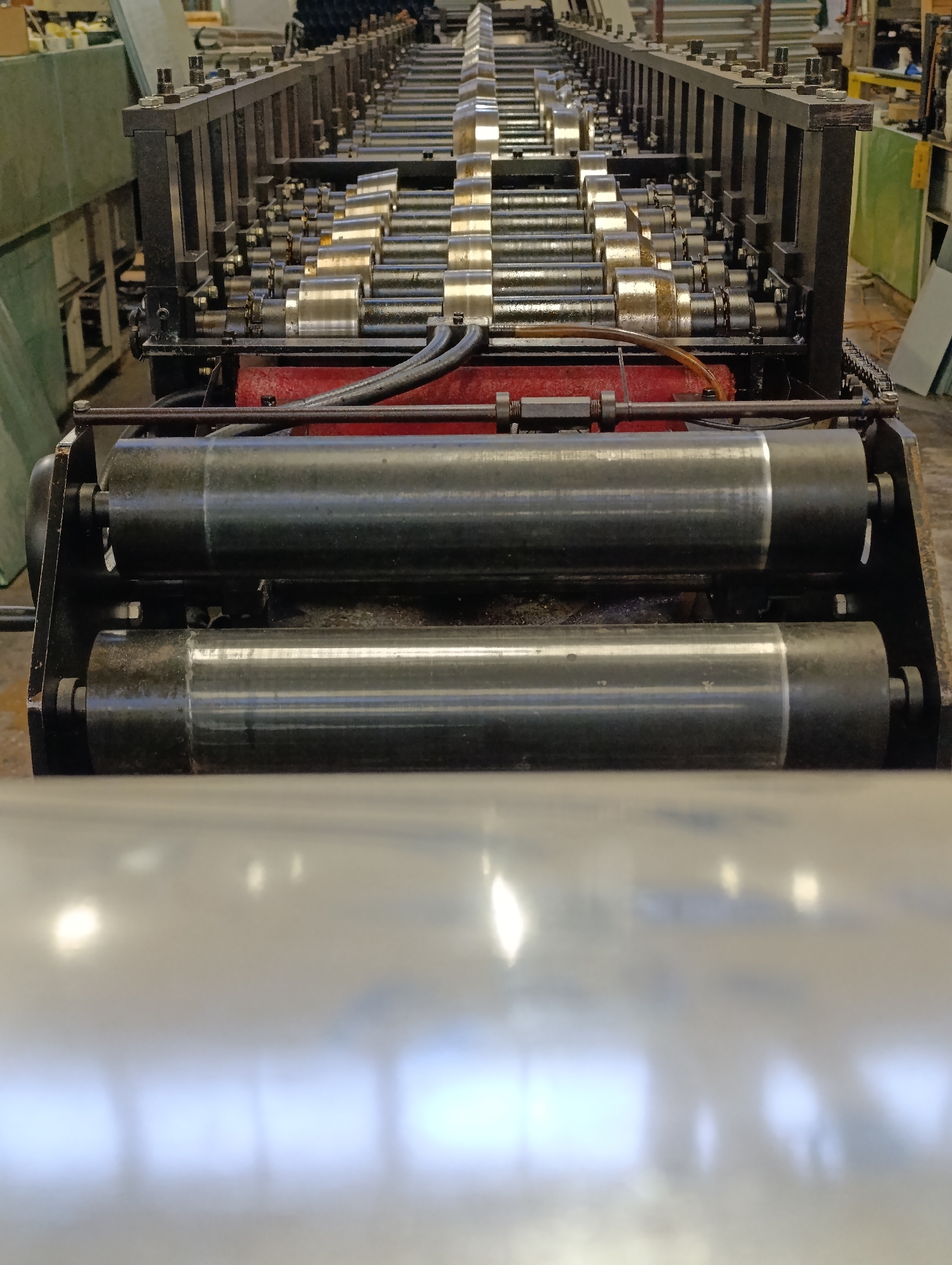 The image shows a metal forming machine in a factory or industrial setting. The machine has numerous rollers and is designed to shape metal sheets. The foreground features a large, shiny metal sheet partially inserted into the machine. The specific location within Haskovo, Bulgaria, is not discernible from the image.