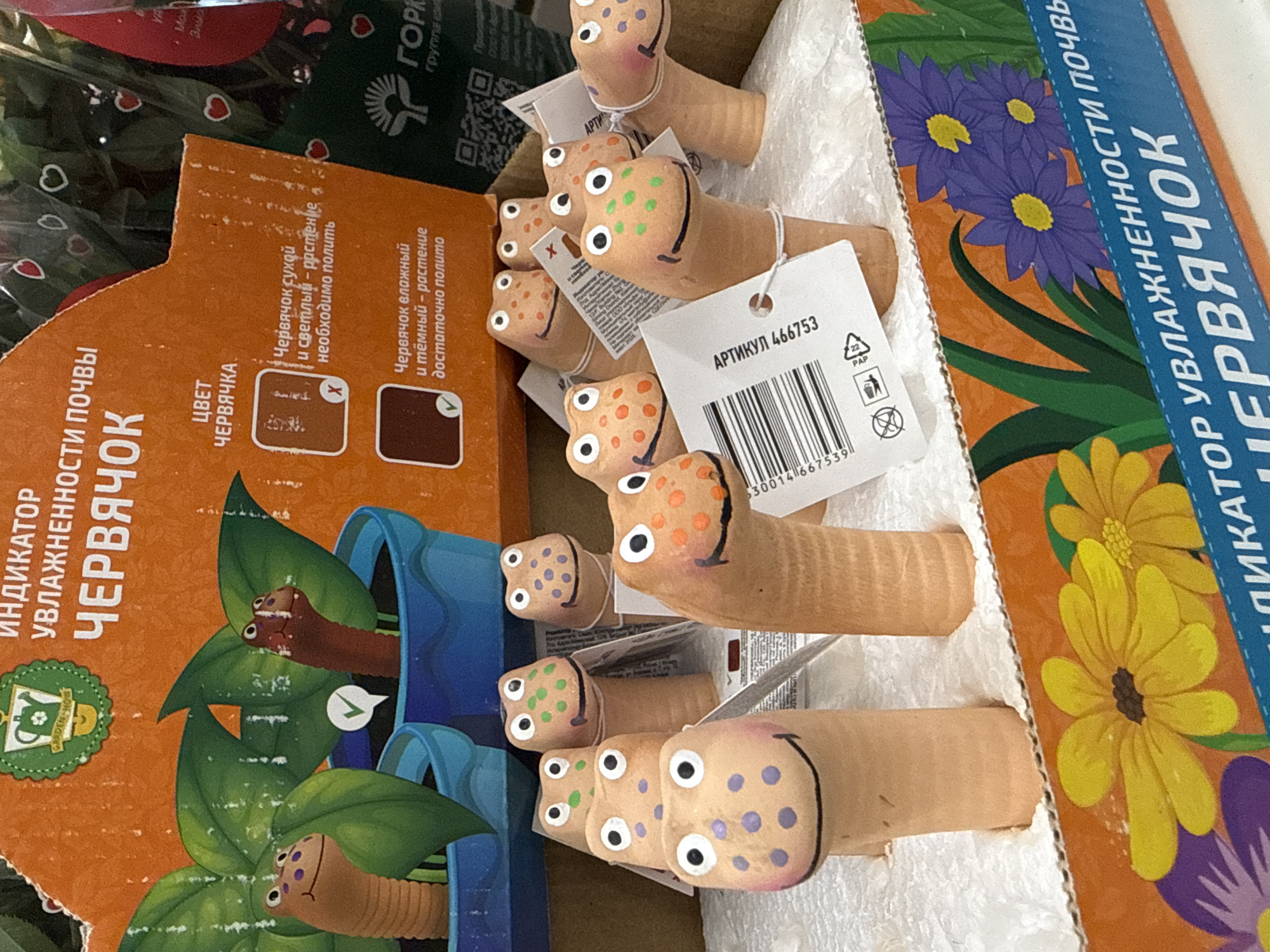 A close-up shot depicts a partially opened cardboard box containing several small, terracotta-colored clay worms. These worm figures are individually tagged with a white label bearing the text "АРТИКУЛ 466753" and a barcode. The head of each worm is adorned with painted spots in various colors, including green, orange, and purple. Adjacent to the box, an orange cardboard package features an image of a worm emerging from a potted plant, along with Russian text that translates to "soil moisture indicator." The package also displays two color samples: one light and one dark. The light color indicates that the plant needs watering, while the dark color indicates that the plant is adequately watered. The scene is indicative of a retail environment in Izhevsk, Russia, likely a garden center or home goods store.