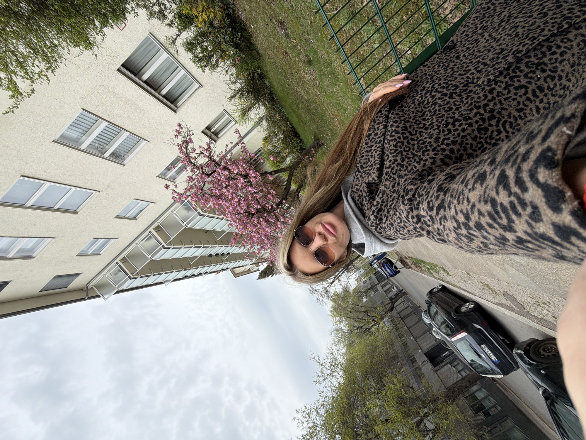 A medium-shot selfie shows a young woman wearing sunglasses and a leopard-print coat. In the background, there is a tall, light-colored building with multiple windows and balconies. A tree with pink blossoms is near the building. A street with parked cars can be seen, and a cloudy sky fills the upper portion of the frame. The location is Berlin, Germany.