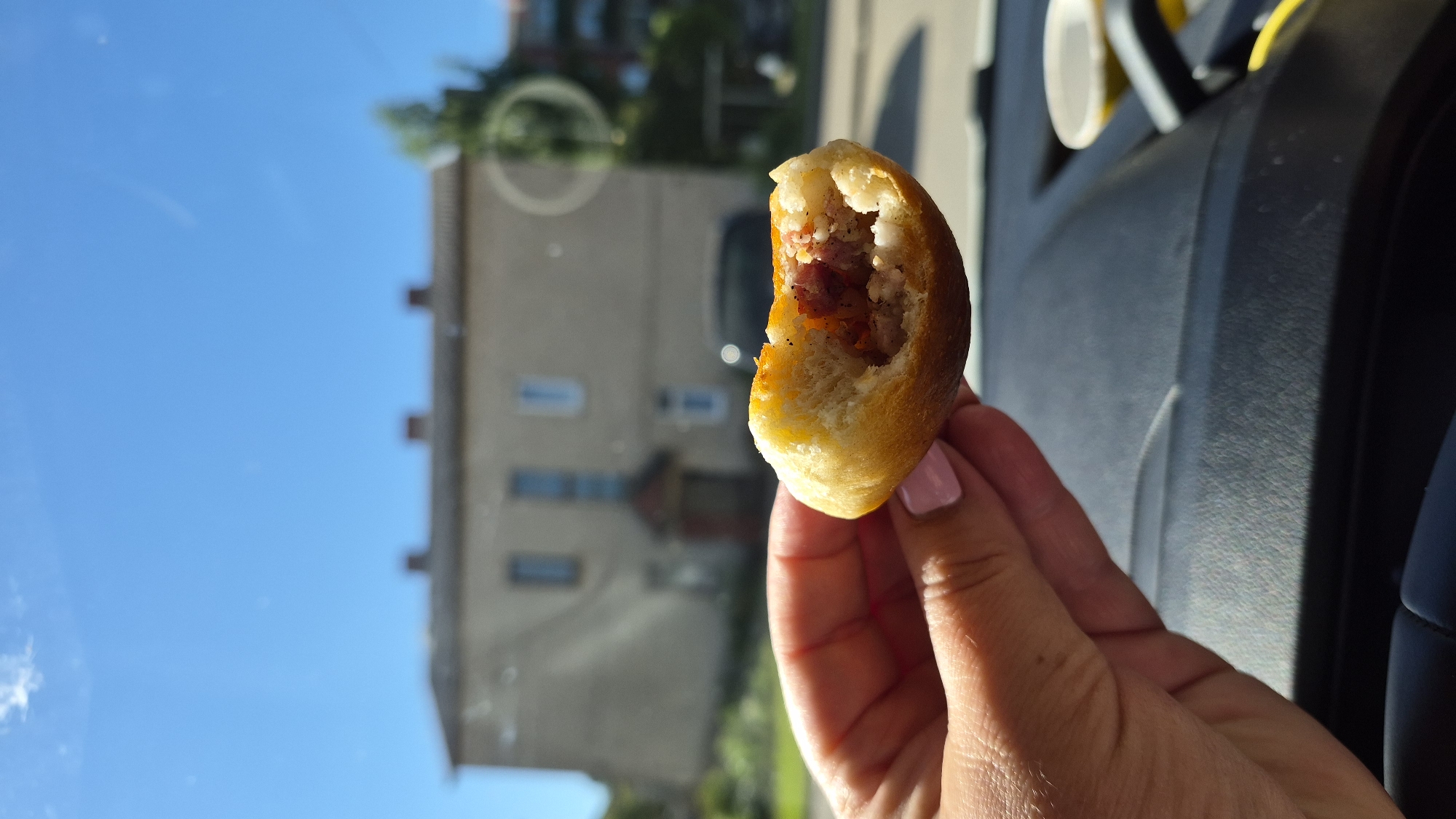 A close-up shot shows a hand holding a partially eaten pastry with a filling visible. The pastry is golden brown and appears to be soft. The hand has light skin with a pink nail polish on one fingernail. In the background, a light-colored building with a dark roof and multiple windows is out of focus. Trees and a clear blue sky are also visible in the background. The foreground shows the dark interior of a vehicle, possibly a car dashboard. A blurred white cup is also visible on the right side of the frame, indicating the scene is inside a vehicle. The setting suggests a casual moment, possibly a snack taken while traveling. The building in the background is a typical residential or small commercial building found in urban or suburban areas. The overall lighting indicates it is daytime with bright sunlight.