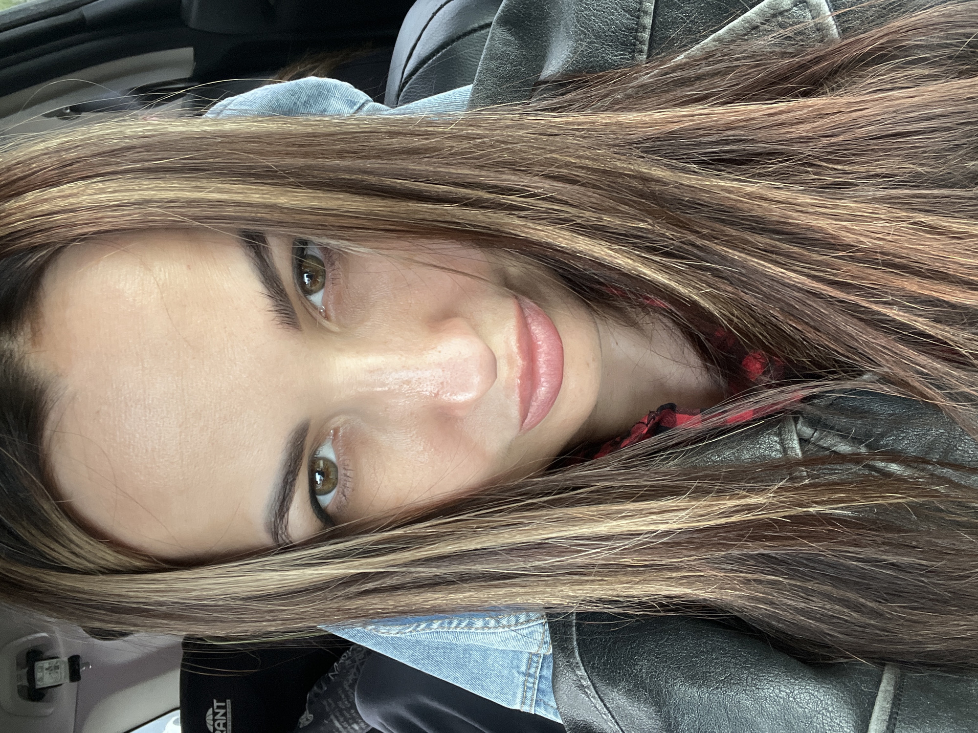 A close-up, head-and-shoulders shot shows a woman with long, light brown hair with blonde highlights, wearing a blue denim jacket over a red and black plaid shirt, and a black leather jacket. Her face is centered in the frame, angled slightly to the left. Her eyes are brown and open, and she is looking directly at the camera with a slight smile. The background is out of focus. The location is Unknown, Unknown.