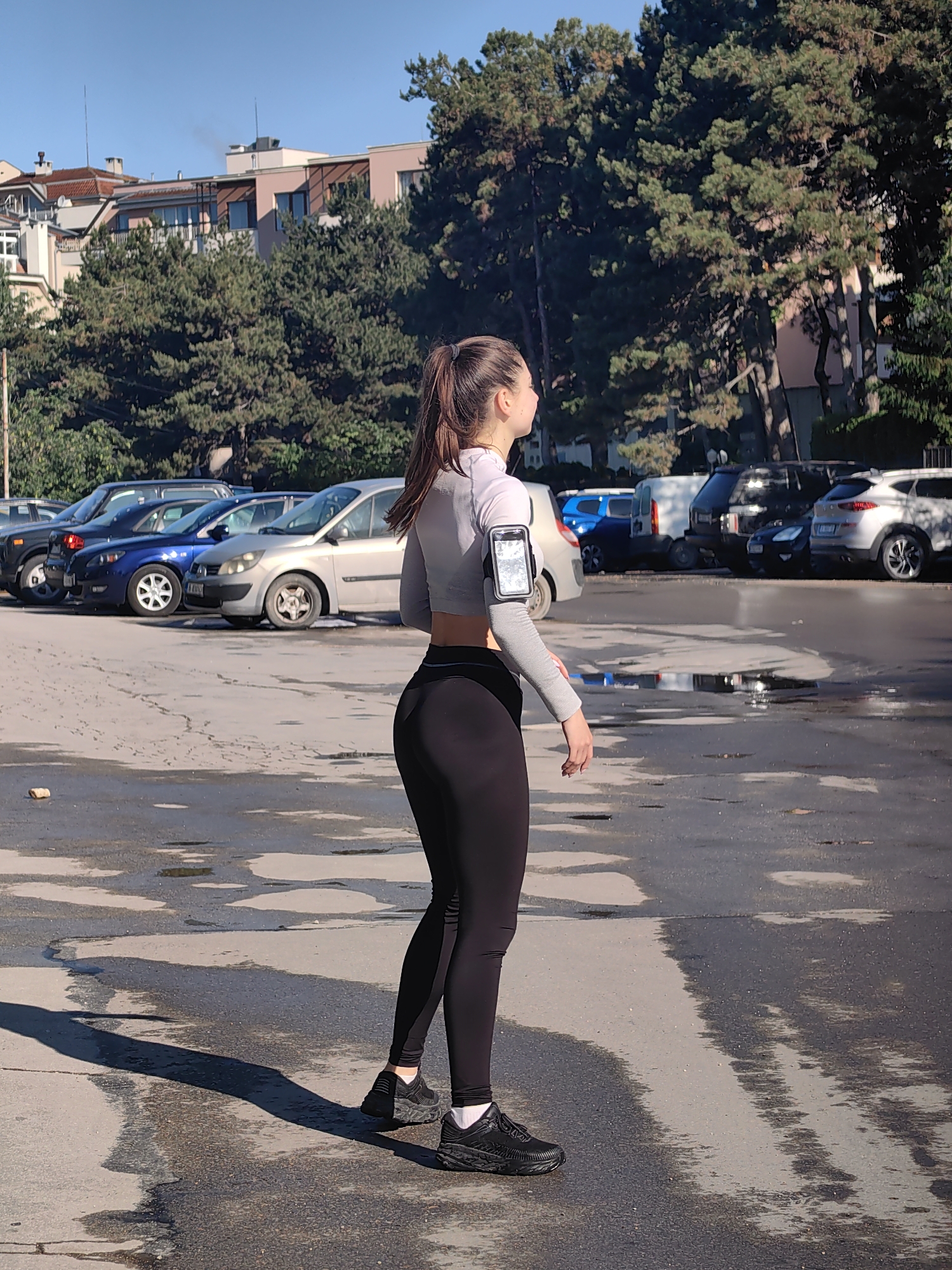 The image shows a young woman in athletic attire standing in a paved lot that appears wet with puddles. She is wearing black leggings, a gray cropped long-sleeved top, and black running shoes. A smartphone is secured to her upper arm with an armband. Her hair is pulled back in a ponytail. She is looking towards the right of the frame.
The lot is bordered by parked cars of various makes and models. In the background, there are buildings and tall green trees.
Based on the architecture and vegetation visible, the location is likely in Varna, Bulgaria. The buildings are reminiscent of urban developments found in many Bulgarian cities, and the type of trees could be seen in Varna's parks and urban areas. The wet pavement suggests recent rain or cleaning.
