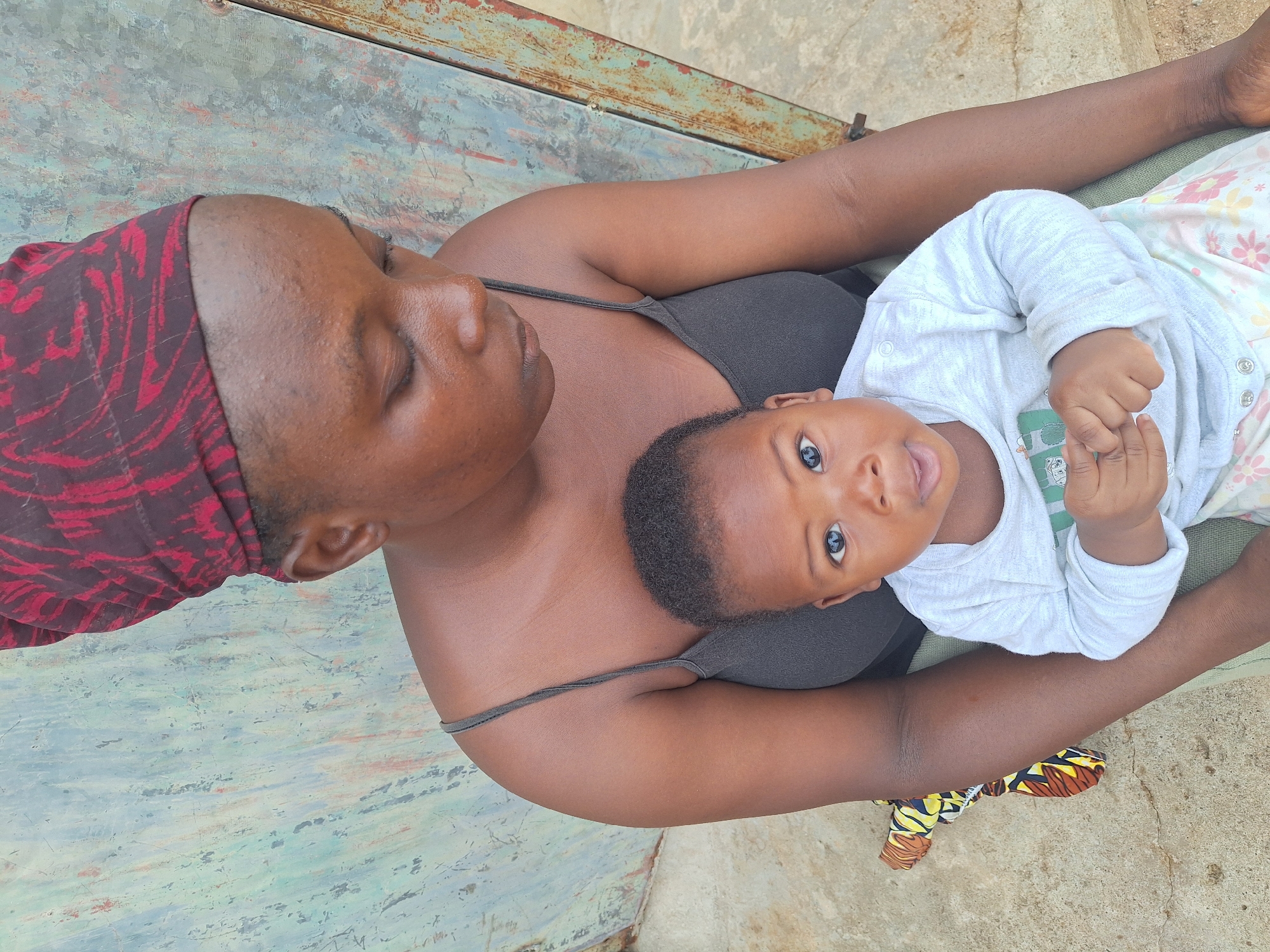 A woman wearing a dark gray tank top and a red and brown patterned head covering is lying on a concrete surface, possibly in the Sabon Tasha region of Nigeria, holding a baby. The baby is wearing a long-sleeved, light gray outfit with printed designs and is looking directly at the camera. The surface they are on appears to be a section of painted metal and concrete. Part of a patterned cloth is visible under the arm of the woman. The woman seems to be in a relaxed position.