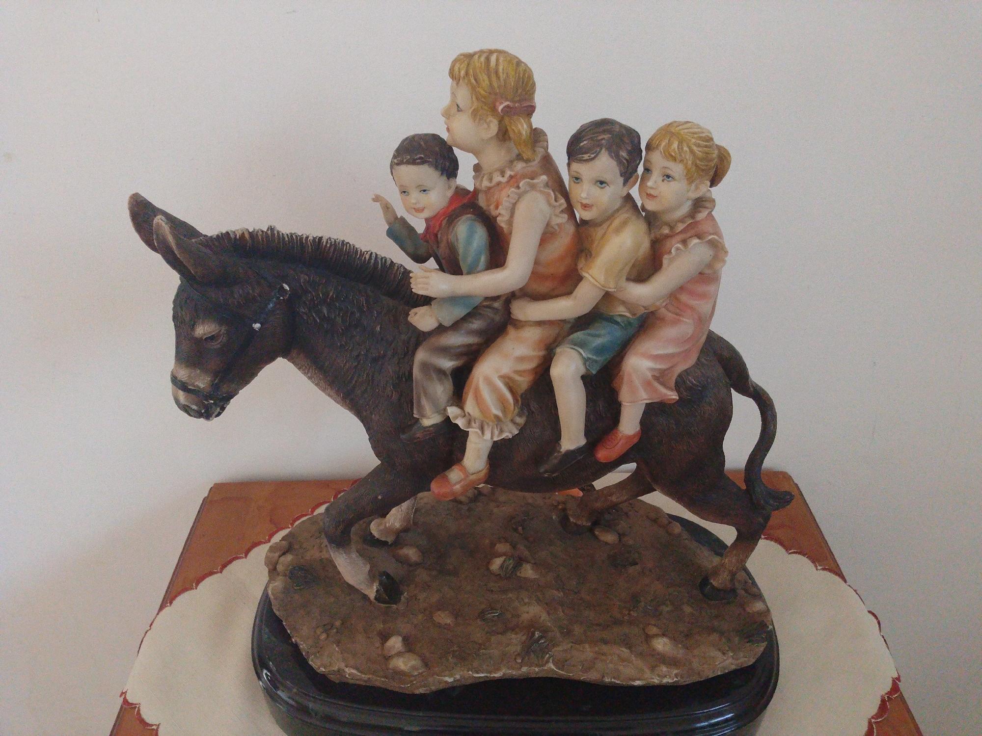 The image shows a figurine of a donkey carrying four children on its back. The donkey is dark brown with a black mane. The children are light-skinned with blonde or brunette hair, and are wearing various pastel-colored clothes and shoes. The figurine is placed on a rough, rock-like base with a dark, glossy, round stand beneath it. The figurine rests on a table covered with a decorative white cloth edged with an orange pattern. The background is a plain white wall. There is no explicit indication of its location in the city of Tinaquillo, Venezuela.