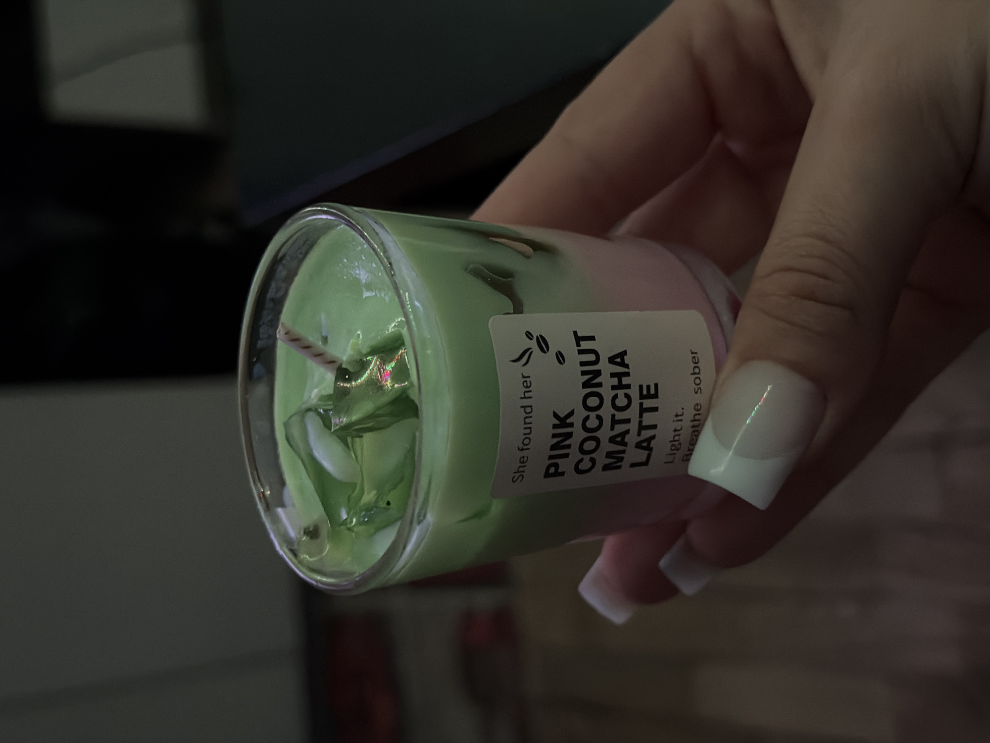 A person is holding a small, clear glass jar containing a layered candle. The top layer of the candle is green, and the bottom layer is pink. Embedded in the green wax is a small, decorative green object that resembles a frog. A label is attached to the jar, displaying the words "PINK COCONUT MATCHA LATTE" in a vertically oriented text. The hand holding the jar has white, square-shaped fingernails with a subtle, light-colored gradient. The background is blurred and features indistinct shapes and shadows. There is no information to indicate this image was taken in Varna, Bulgaria.