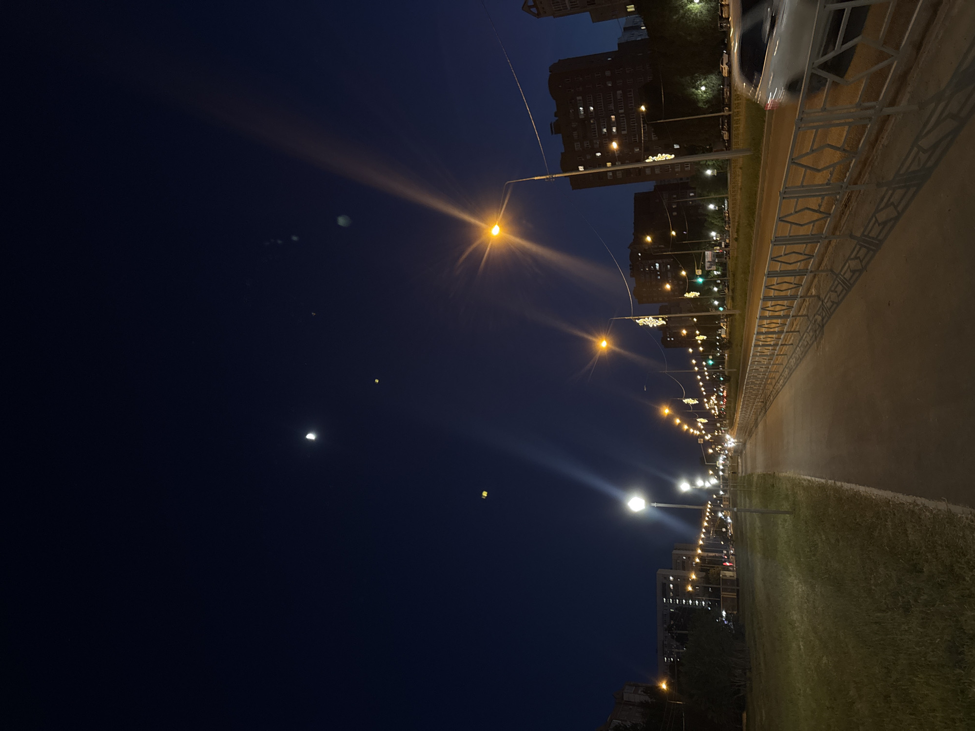 The image depicts a night scene in an urban environment. The sky is a deep blue, with a visible crescent moon. Streetlights line the right side of the image, illuminating the road and surrounding buildings. A metal railing runs along the edge of the road, separating it from a grassy area. Buildings are visible in the background, their windows lit up. The photograph is taken from a slightly elevated position, looking down the street. There are no people visible in the scene. The overall impression is one of a quiet, well-lit urban street at night.