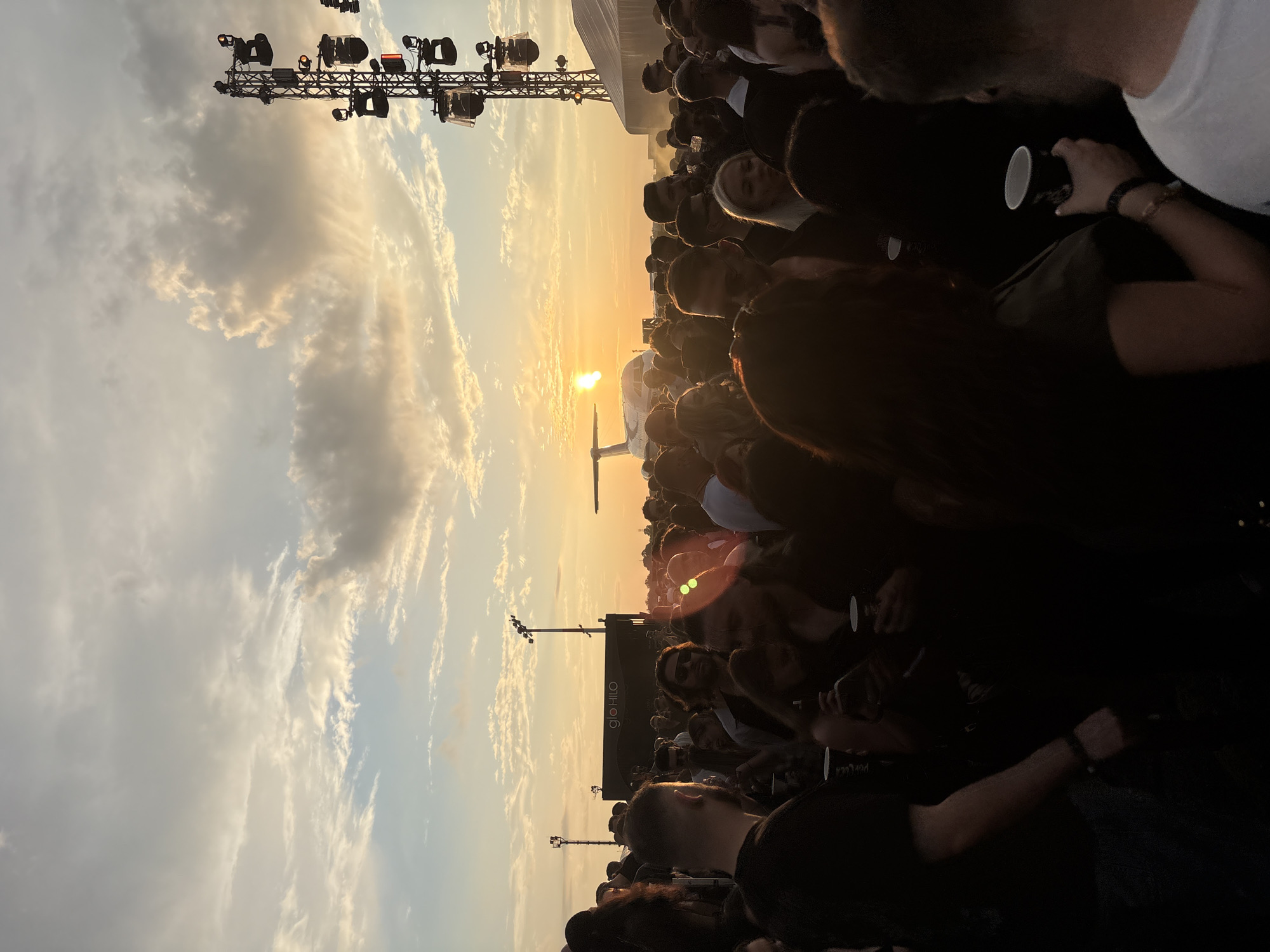 The image depicts a crowded outdoor event at sunset, likely an airfield, identified as Romaero Băneasa in Bucharest, Romania. The scene is dominated by a large gathering of people, mostly young adults, standing and mingling under a partly cloudy sky illuminated by the low, setting sun.
In the mid-ground, a white, narrow-body jet aircraft is prominently visible, silhouetted against the bright orange and yellow hues of the sunset. A significant sun flare emanates from the vicinity of the aircraft's left wing and engine, obscuring some detail. The plane appears to be parked on a tarmac or open field adjacent to the event area.
On the far right, a tall metal truss structure, typical of concert staging, supports multiple stage lights and speakers. Some of these lights emit an amber glow. To the left, behind the crowd, a large black rectangular structure bears the white "glo HILO" branding. Part of a large white tent structure is also visible on the far right.
The crowd fills the lower two-thirds of the frame. Many individuals are silhouetted, with faces and details obscured by the backlighting. Several people in the foreground and mid-ground are holding disposable cups. The overall atmosphere suggests a music festival or similar large-scale public event.