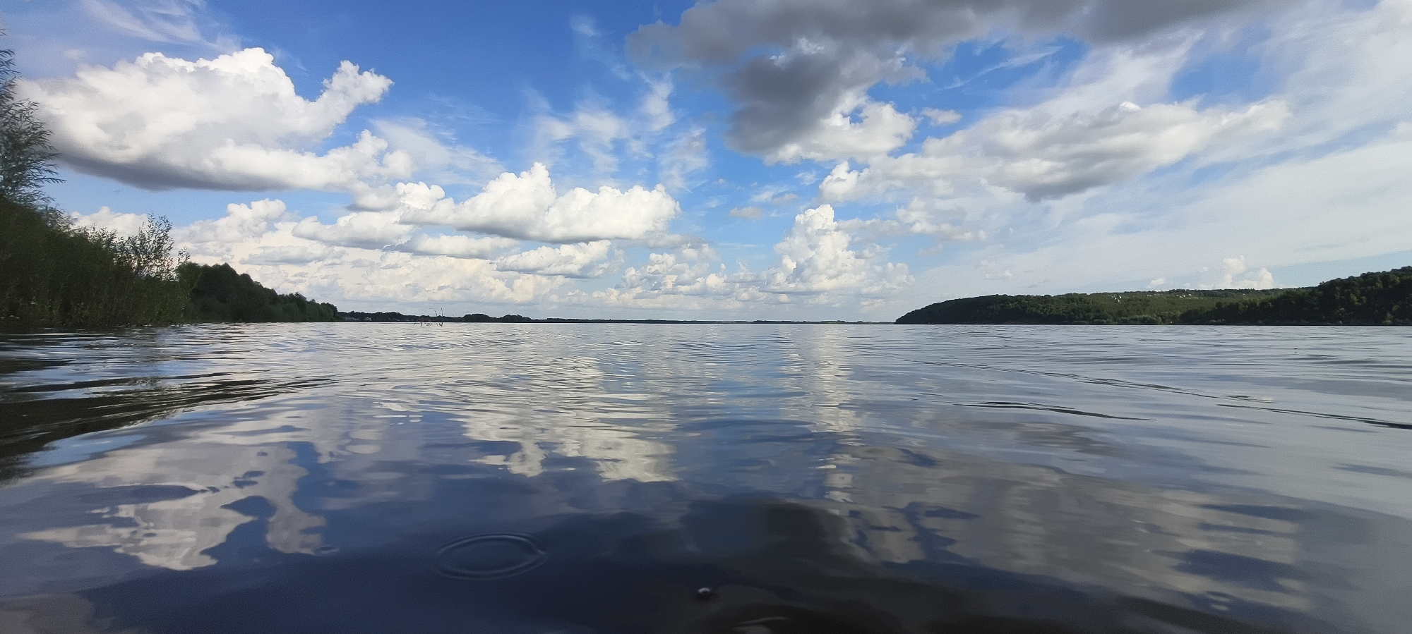 The image captures a wide panoramic view of a calm lake under a bright blue sky filled with fluffy white and dark gray clouds. The foreground is dominated by the rippling surface of the water, reflecting the sky and clouds. On the left side of the frame, a lush green shoreline with dense trees and bushes is visible. In the distance, the opposite bank of the lake features a rolling, tree-covered hillside. The overall scene depicts a serene natural landscape on a partly cloudy day.