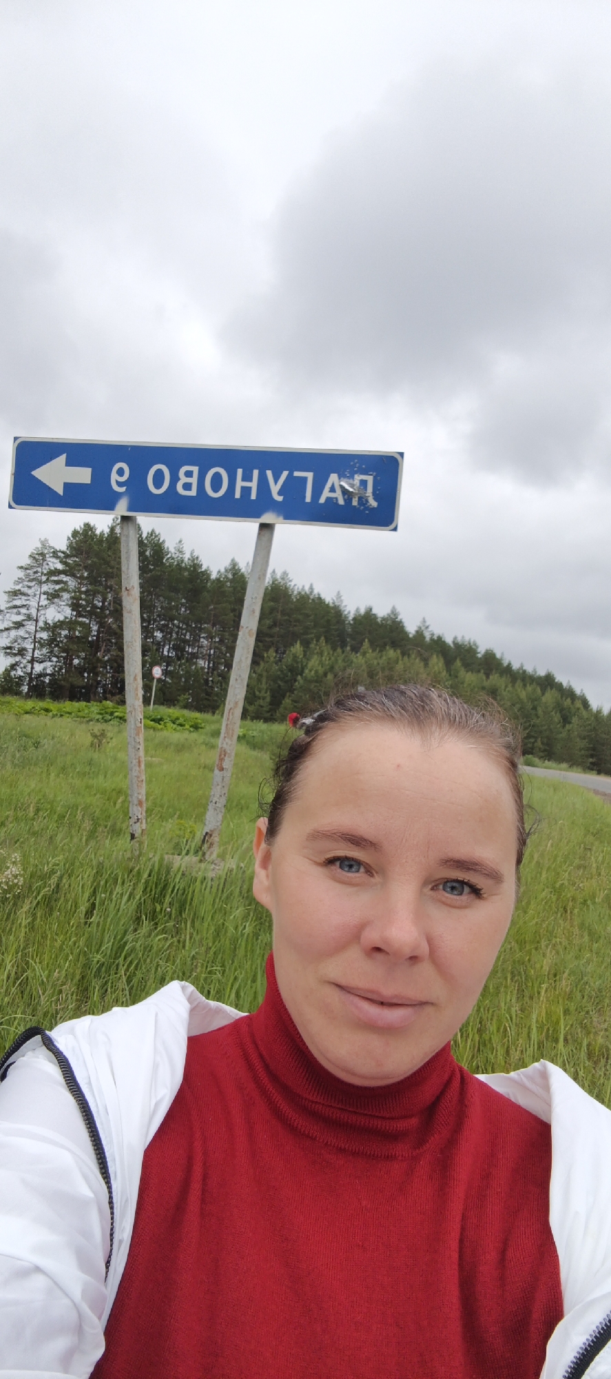 A frontal outdoor medium shot features a woman in the foreground with a road sign and natural landscape in the background under an overcast sky. The woman, positioned centrally, has fair skin, blue eyes, and dark hair pulled back with some gray strands, adorned with small red hair accessories. She wears a red turtleneck top and a white jacket with a black zipper, draped over her shoulders, and exhibits a subtle smile while looking directly at the camera. Behind her, a blue rectangular road sign is visible, mounted on two weathered gray poles. The sign displays a white left-pointing arrow and mirrored white Cyrillic text, which reads "е ОВОНУЛГАД" when read left to right as seen in the reflection. When un-mirrored, the text likely spells "ГАЛУНОВО е" (GALUNOVO e). Behind the sign and woman, tall green grass fills the immediate background, leading to a dense forest of pine trees stretching horizontally across the midground. A small white circular sign, possibly a traffic sign, is partially visible behind the right pole of the blue sign. A paved road is discernible on the far right. The sky is largely covered by uniform gray clouds. The location is unknown.