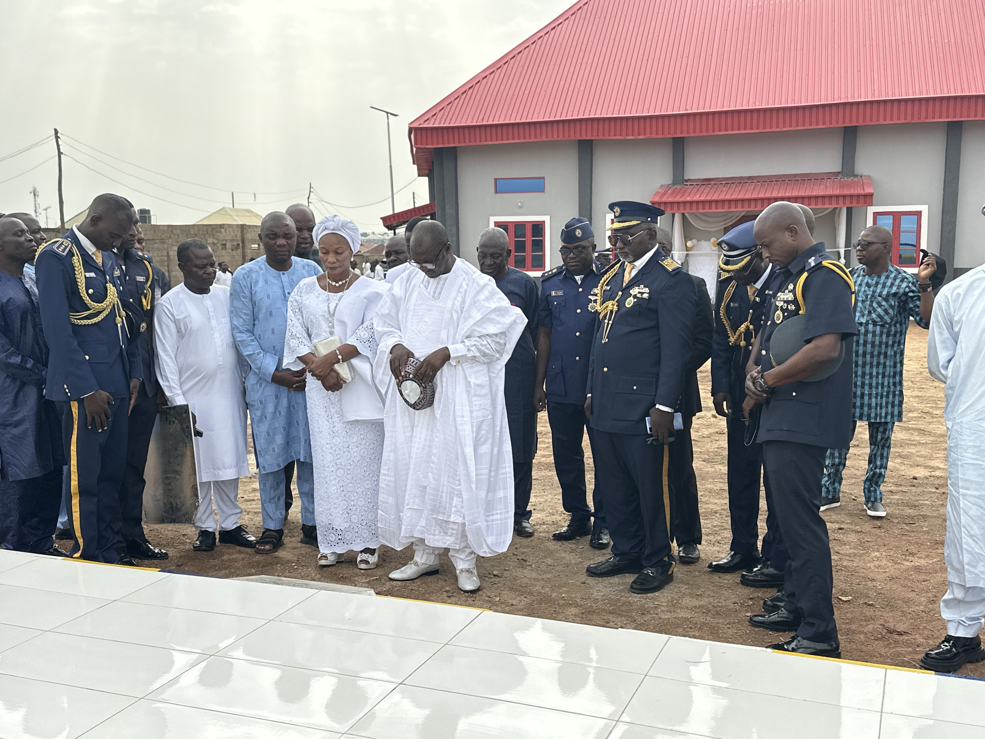 In Goni Gara, Nigeria, a group of people are standing together outside a building with a red roof and light gray walls. The group includes men and women dressed in a variety of clothing styles. Some of the men are wearing military-style uniforms with gold accents, while others are dressed in traditional Nigerian attire, including white robes. One woman is wearing a white lace dress and a headwrap. The people are looking down at a white tiled surface. The sky is overcast. The ground is dirt.