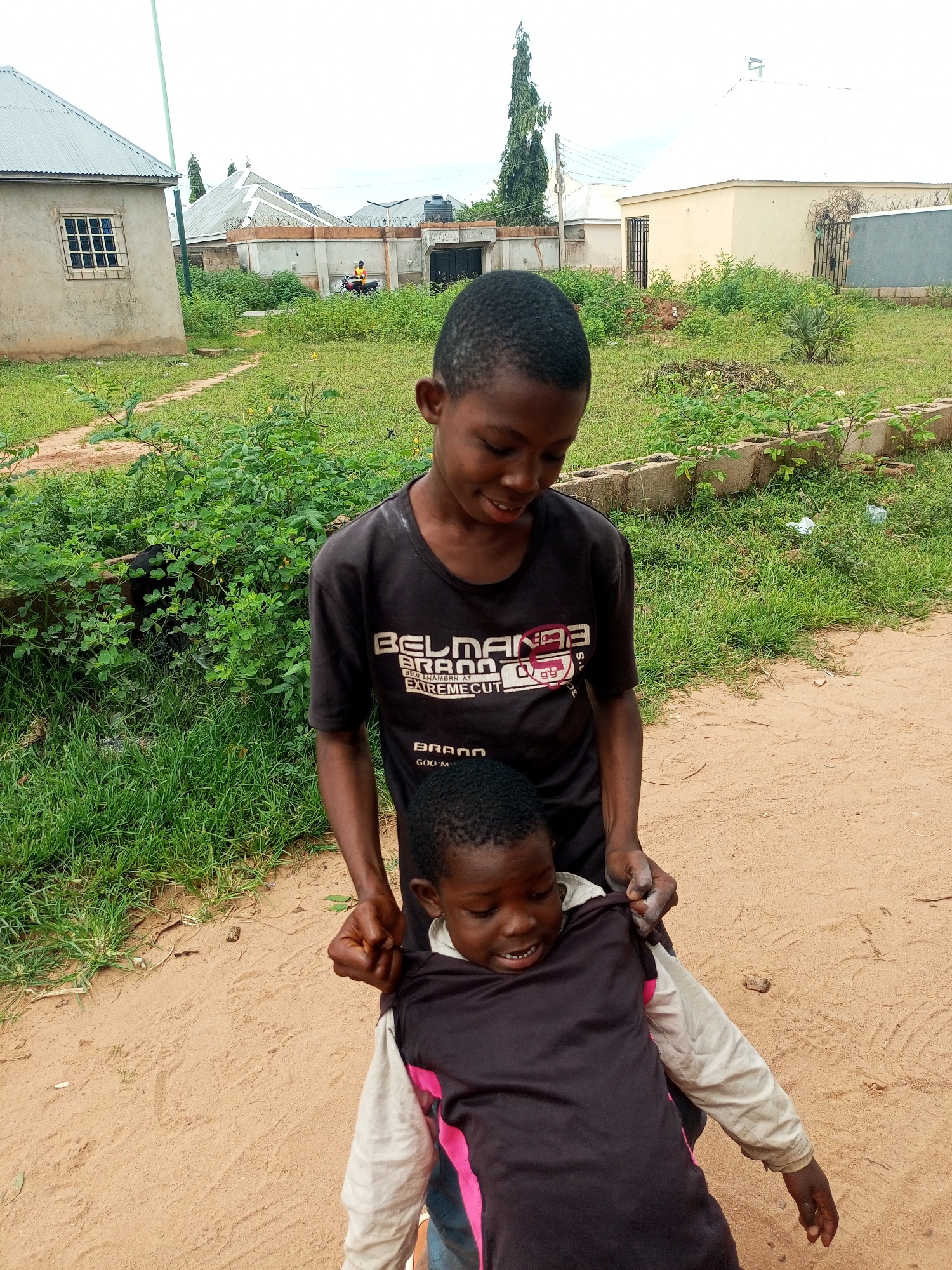 In Tempure, Nigeria, two young boys are interacting on a dirt path beside lush green vegetation. The older boy, wearing a black t-shirt with white lettering that reads "BELMANBR AT EXTREMEECUT", stands behind and to the side of the younger boy, his hands gripping the younger boy's shirt. The younger boy, facing away from the camera, is wearing a dark long-sleeved shirt with pink accents on the sleeves and a lighter colored shirt underneath. The background reveals a residential area with a light-colored house with a metal roof, a fence, and more vegetation. In the distance, another house with a cream-colored exterior and a light-colored roof is visible, along with a tall, thin tree. The sky is overcast.
