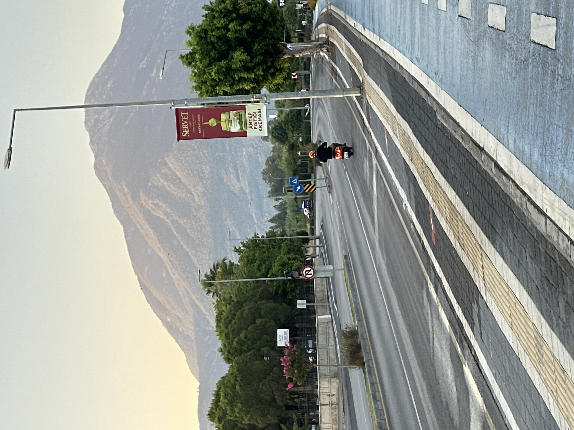 The image depicts a street scene in Fethiye, Turkey, at what appears to be dusk or dawn, given the soft, diffused light. Dominating the background is a large, hazy mountain range. In the middle ground, a paved road curves to the right, with a dedicated lane marked by a blue surface on the right side. A single motorcyclist wearing a red helmet and a dark jacket is riding away from the viewer on the road. Adjacent to the road is a pedestrian walkway with tactile paving. A tall street lamp stands prominently on the right, with a vertical banner attached to it advertising "SERVET" and "ANTEP FISTIĞI KREMASI" (Pistachio Cream). Several trees with dense green foliage are visible along the roadside. Further down the road, a police car with flashing lights is parked. Traffic signals and road signs are also present. The overall environment suggests an urban or semi-urban setting.