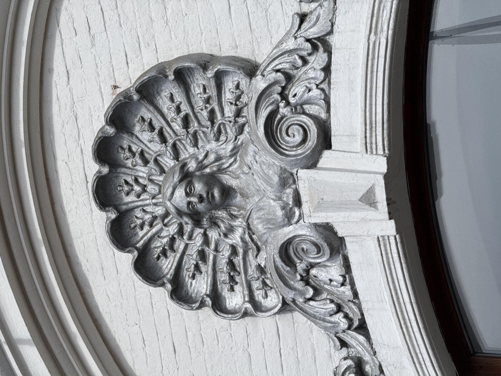 Close-up shot of architectural detail featuring a face in a floral motif, possibly an allegorical representation, mounted on a white brick wall. To the right, decorative column design elements are visible, next to a curved window frame. The metallic-grey color could imply the material used is bronze or painted metal. Exact city unknown.