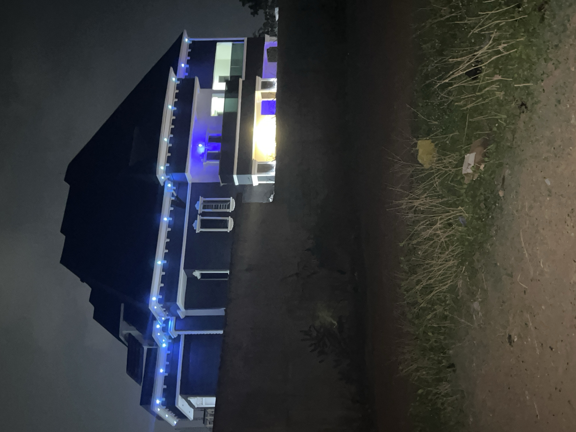The image shows a multi-story building at night, likely a residential property, with a sharply angled roof and light-colored trim. The building is illuminated with white and blue lights, and light emanates from some of the windows. The surrounding environment is dark, with a patch of grass and a dirt road or path visible in the foreground. There are no people visible in the image. While the location in Abuja, Nigeria is provided, there are no specific attributes in the image to confirm the place. The scene is quiet and still.
