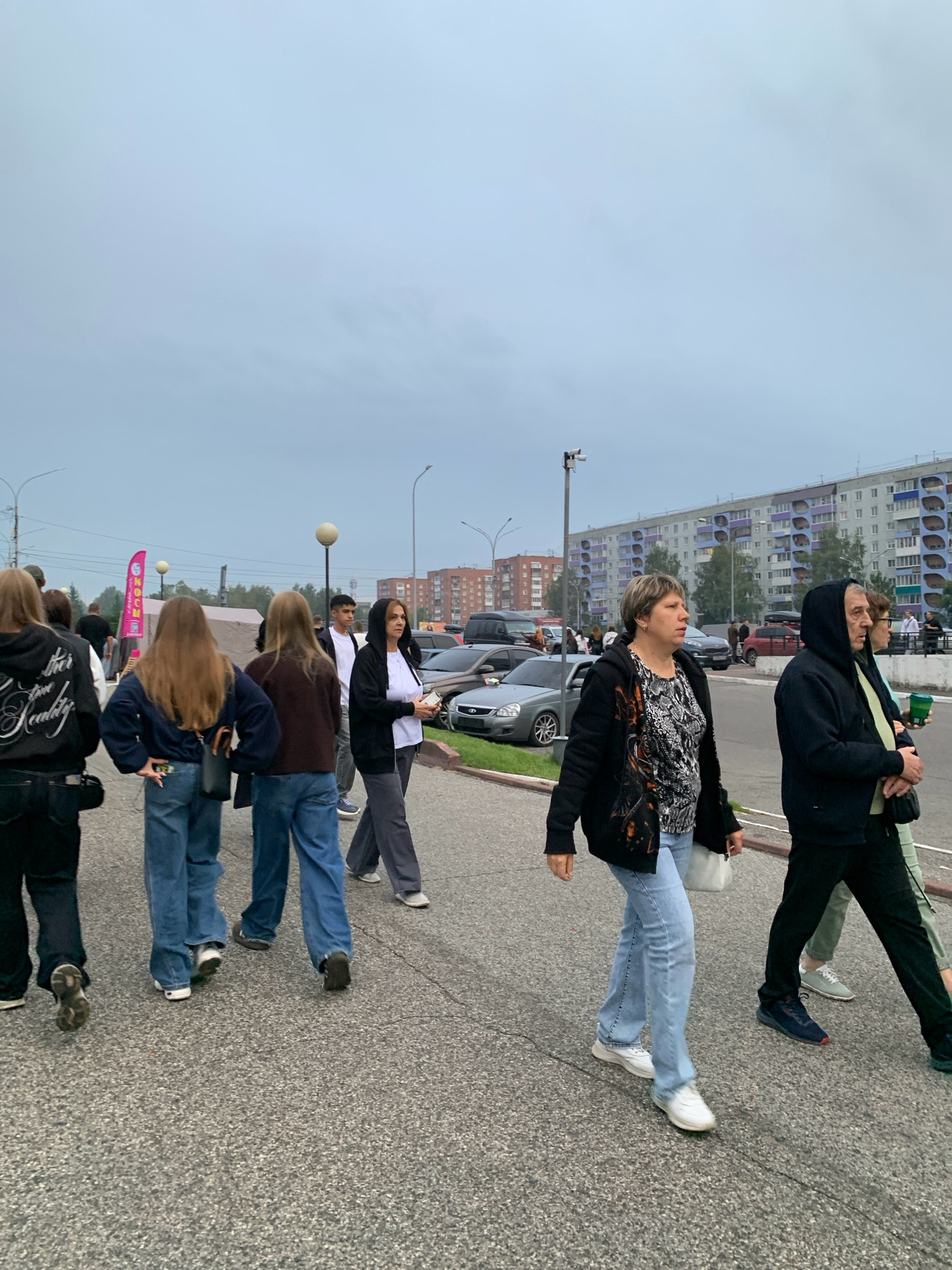 A group of people are walking on a paved sidewalk in front of apartment buildings and parked cars. The sky is overcast. A pink banner with Russian text is visible in the background. The scene is likely in Prokopyevsk, Russia, based on the provided information about the city.