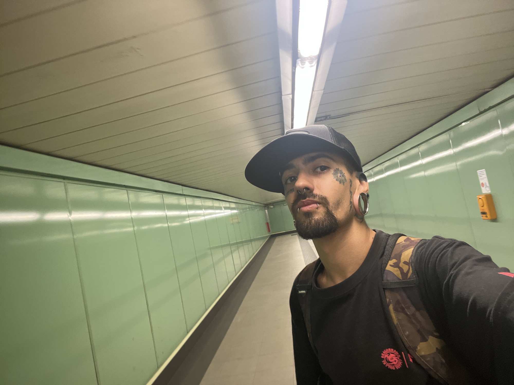 The image shows a person wearing a black baseball cap and a black long-sleeved shirt with a camouflage backpack. The person has a tattoo on their cheek, a nose ring, and stretched earlobes. They are standing in a subway station with green walls and a long corridor. The ceiling has fluorescent lights. There is an orange button on the wall to the right of the person. The overall environment suggests a public transportation area. The location is identified as a subway station in Madrid, Spain.