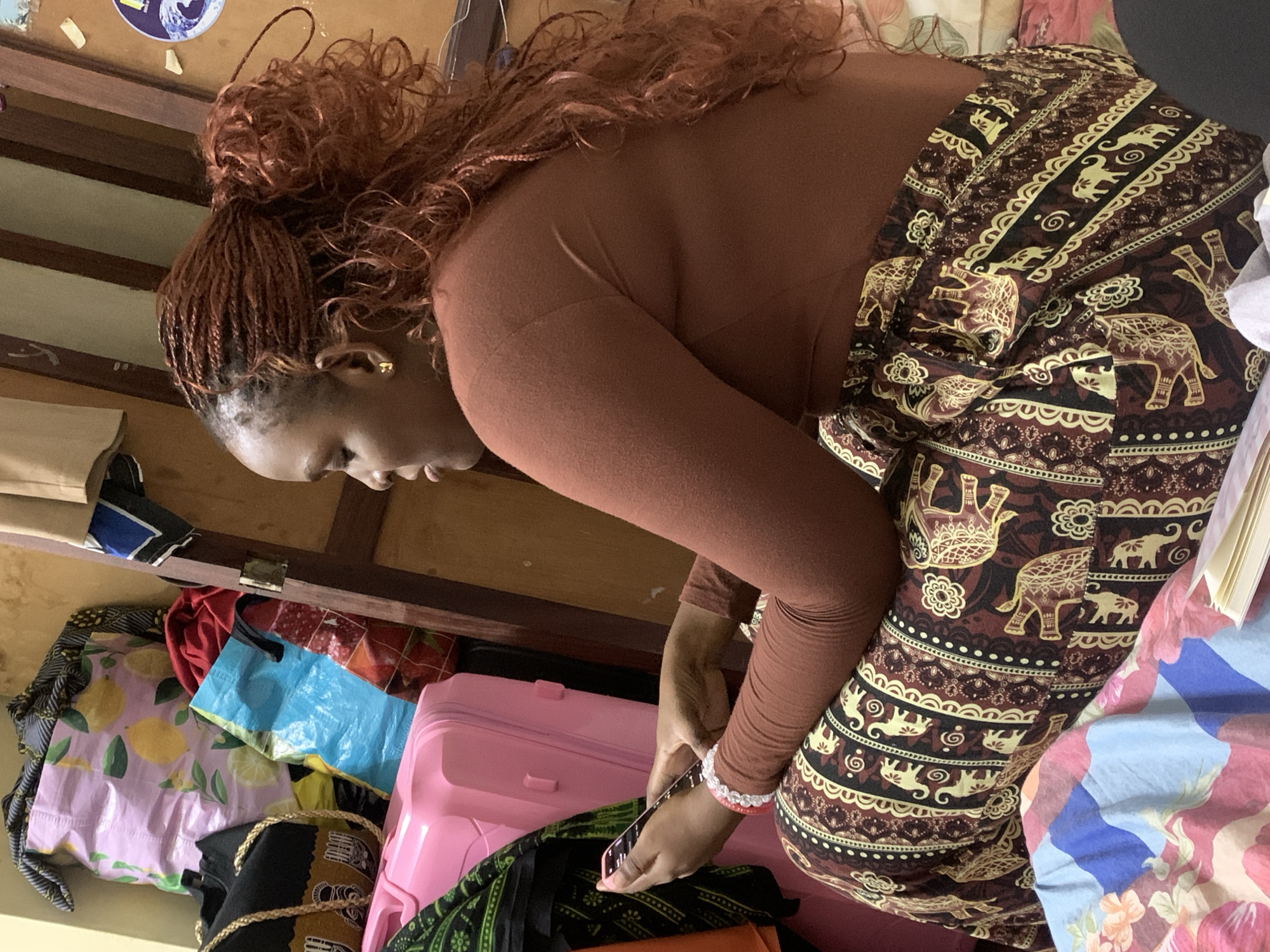 A woman is seated in profile, facing left, against a light brown wall in what appears to be an indoor residential or temporary accommodation setting. She wears a long-sleeved brown top and a dark brown fabric wrap (likely a kanga or kitenge) patterned with light brown/cream elephants and floral motifs. Her hair is styled in reddish-brown micro braids, pulled back into a high, curly ponytail. A small gold-colored earring is visible in her right ear, and a white and pink beaded bracelet is on her left wrist. She is holding a smartphone with both hands, appearing to interact with it.
To her left, a pink hard-shell suitcase stands upright, partially obscured by a draped dark green patterned fabric. Behind and above the suitcase, a stack of items includes a light purple reusable shopping bag with a lemon pattern, a blue plastic bag, and a red fabric bag. A wooden frame, possibly part of a door or furniture, runs vertically behind her, with a circular blue and white sticker affixed to the wall or frame above her shoulder. The woman is seated on a surface covered by a light-colored patterned fabric, featuring blue, pink, and white floral and geometric designs. A small, open book or notepad is partially visible on the surface near her right leg.
The specific location within the city of Dar es-Salaam, Tanzania, cannot be determined from the visual information provided.