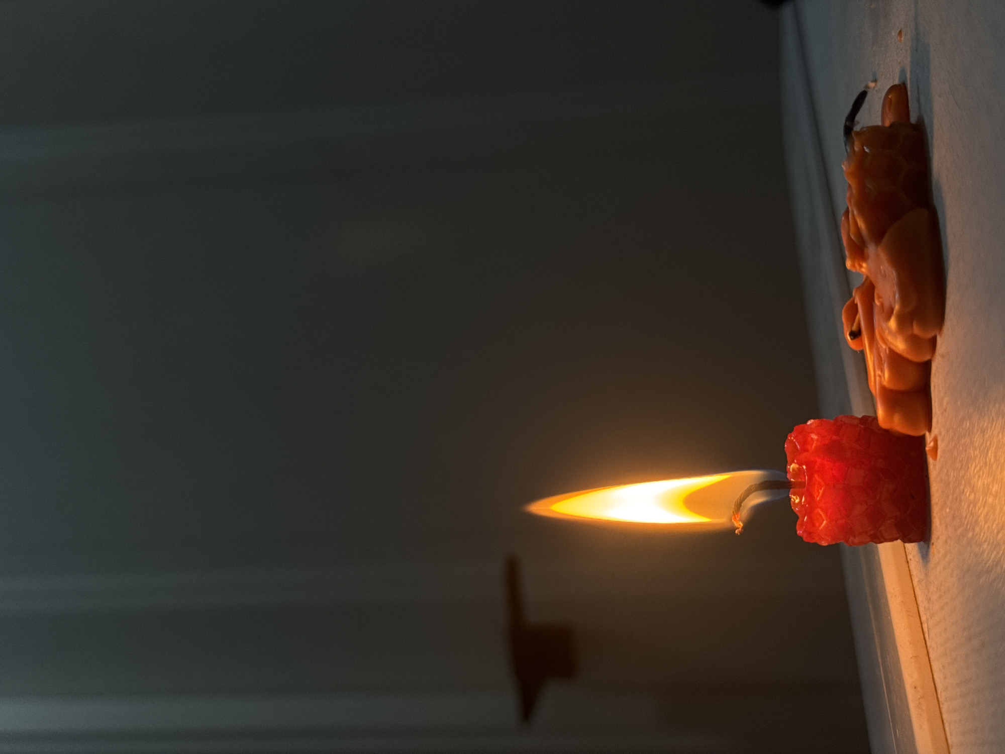 A side view shows a red candle with its wick burning on a white surface. Next to the candle is hardened, dripping, orange candle wax. The background is dark with vertical gradations in grayscale. The location is Unknown, Unknown.