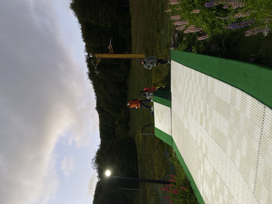 The scene presents a synthetic ski slope nestled amongst lush greenery. Two people, dressed in casual attire, stand near the platform, possibly observing or instructing. A ski lift structure reaches into the background, hinting at winter activities. Lupines bloom near the top, indicating a vibrant summer environment. The light pole stands as a testament to human intervention. Above, the sky is covered with dark clouds, with some sky visible in between.