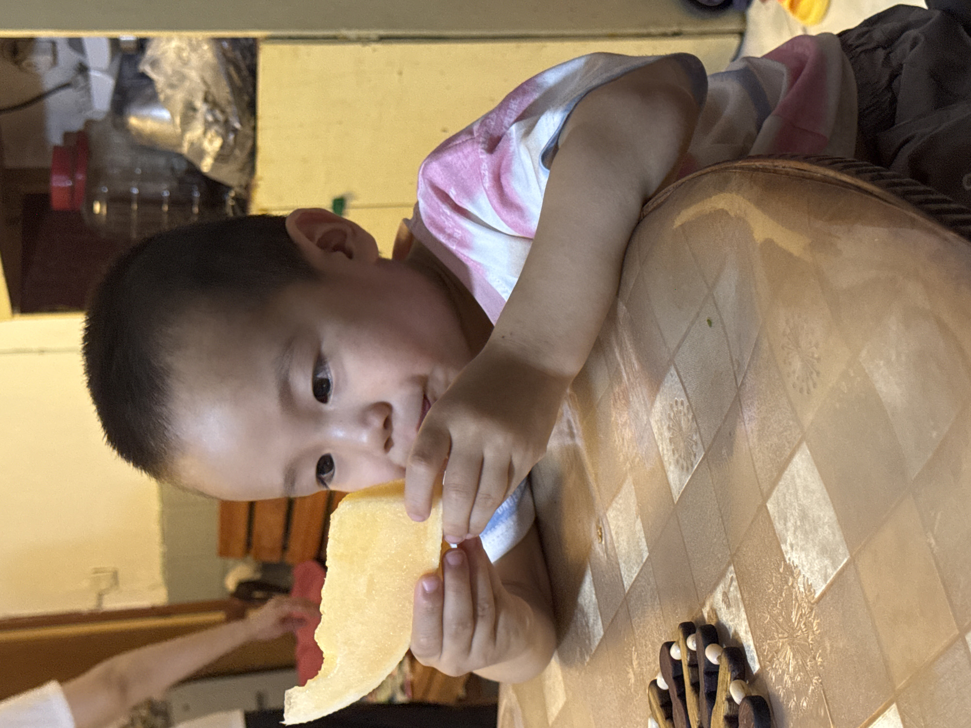 An overhead, close-up shot shows a young East Asian child, likely a boy, sitting at a round, brown table in what appears to be a home. The child is holding a slice of cantaloupe up to their mouth with both hands, as if about to take a bite. They are wearing a tie-dye t-shirt with pink and white stripes, and dark pants. The table has a checkered pattern with some floral motifs. To the left of the child are some wooden game pieces. In the background, on a shelf, there are jars, bags of food, and other household items. A person is partially visible on the left edge of the frame, with their arm extended. The scene is brightly lit, suggesting daytime. The location is identified as Guandu, China.