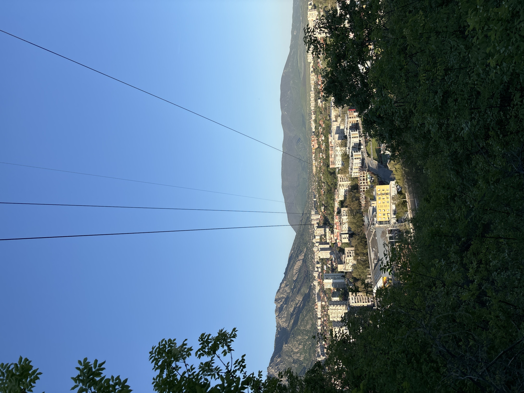 The image presents a view of a city nestled along a mountain range, potentially from an elevated position. The sky is a clear blue, with a set of power lines stretching across the upper left portion of the frame. A city unfolds on the right side, at the base of the mountain. Buildings of various sizes and architectural styles dot the landscape, with streets and possibly some green spaces visible between them. Trees are visible in the foreground.