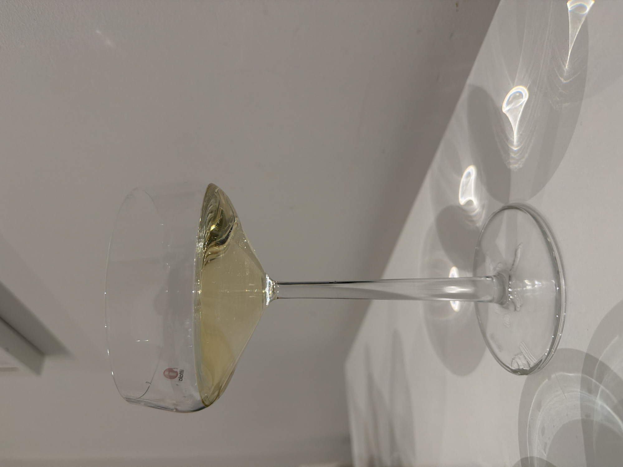 A glass of white wine rests on a white surface, likely a table. The glass is partially filled, and light from an unseen source creates reflections on the table around the base of the glass. The presence of the “blotfii” logo suggests the glass is a product of the Blotfii brand. There is no indication of the location being in Berlin, Germany.
