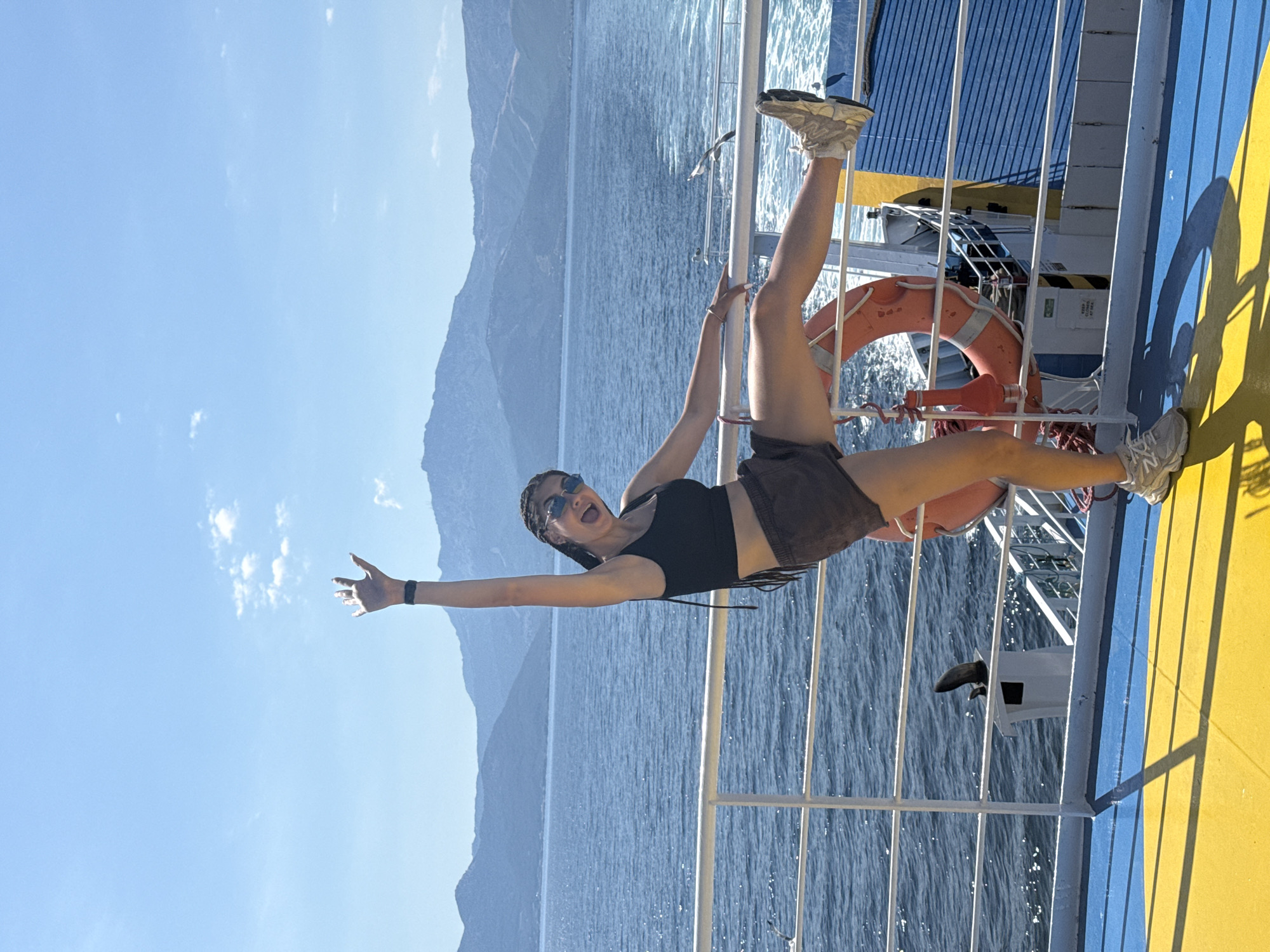 A young woman with braided hair and sunglasses is posing playfully on the deck of a ferry. She is wearing a black crop top and brown shorts. Her right arm is raised in the air, and her left leg is extended outwards. Behind her, the blue sea stretches out to a mountainous coastline under a clear sky. The ferry deck is painted with yellow and blue stripes. A life preserver is visible on the railing.