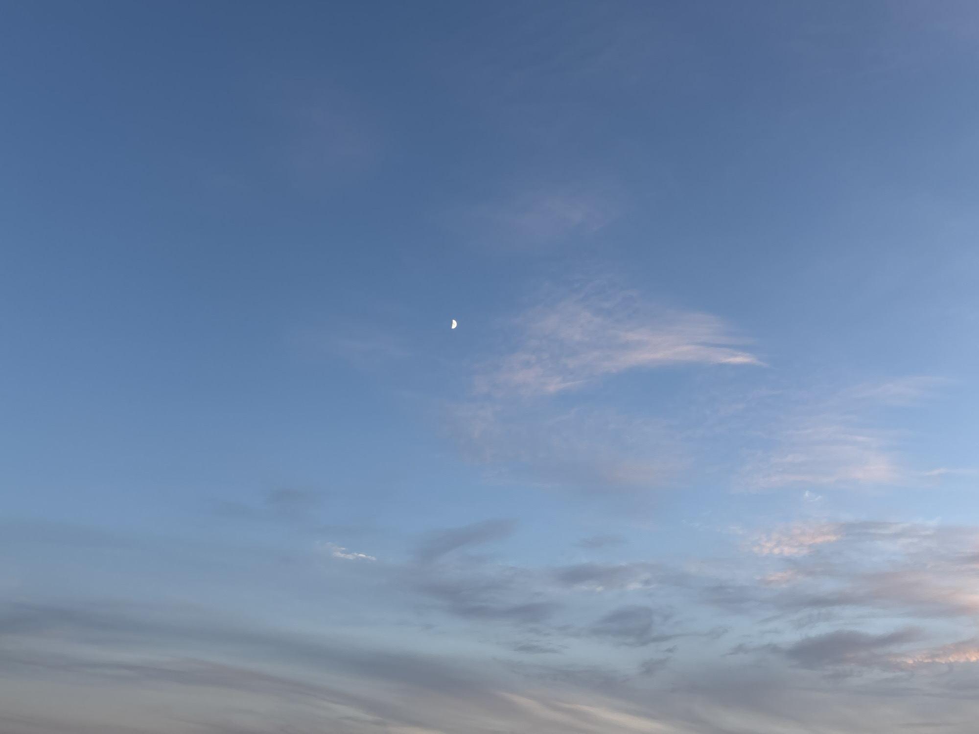 The image shows a clear sky with a crescent moon visible in the upper left quadrant. Wispy clouds, tinged with pink and orange hues, are scattered across the sky, particularly in the lower half of the frame. The lower part of the image is dominated by these clouds, which transition into darker shades of grey and white towards the horizon. The overall lighting suggests it is either sunrise or sunset. There are no people, buildings, or any other identifiable objects or activities present in the scene. The image is an outdoor, atmospheric photograph of the sky.