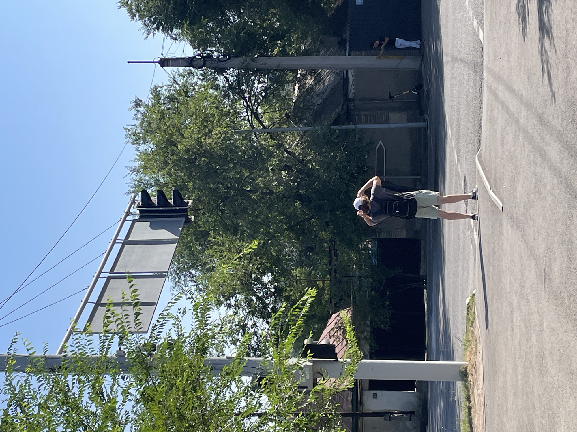 A young male with a backpack and light-colored shorts stands on a street corner in Almaty, Kazakhstan, facing away from the camera and adjusting his white cap. He is near a traffic light. To his right, a person in a black shirt and white pants is near a large dark gate, and a yellow scooter is parked nearby. The street appears to be an intersection with painted white lines. Trees with dense green foliage are visible throughout the scene, and a clear blue sky is overhead. The buildings in the background are simple, with tiled roofs and a somewhat rustic appearance. The image captures a typical sunny day in an urban environment.