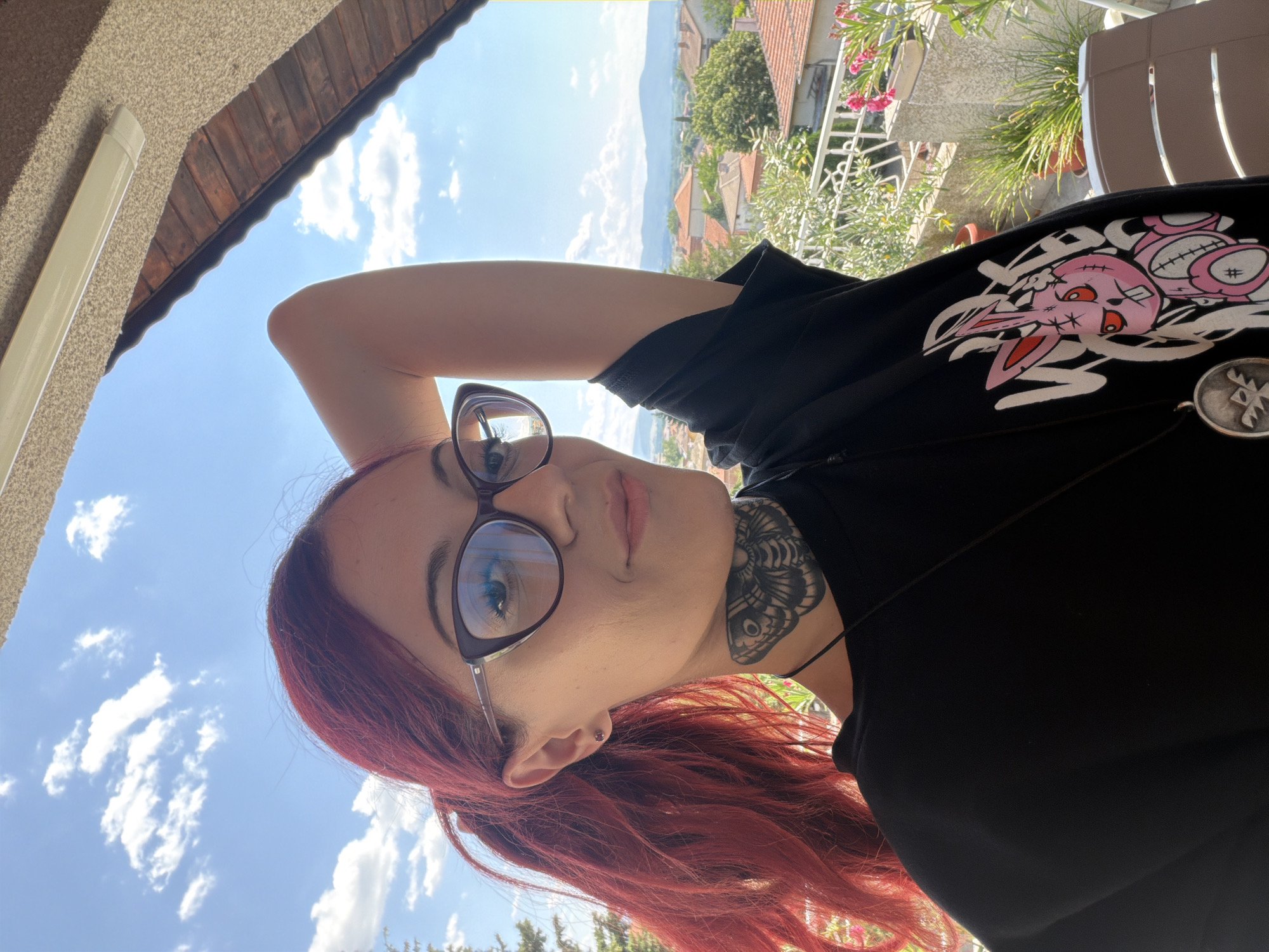 The image shows a young woman with bright red hair, wearing glasses, a black t-shirt with a pink graphic of a bunny, and a necklace. She has a butterfly tattoo on her neck. She is standing on a balcony overlooking a city with red-tiled roofs and distant mountains, likely Svilengrad, Bulgaria, under a clear blue sky with scattered clouds. The woman is holding her arm up behind her head, and the camera is positioned at a slight upward angle, creating a selfie-style portrait. Plants and pots are visible on the balcony, along with a chair. The lighting suggests it is daytime, possibly late morning or afternoon, with bright sunlight.