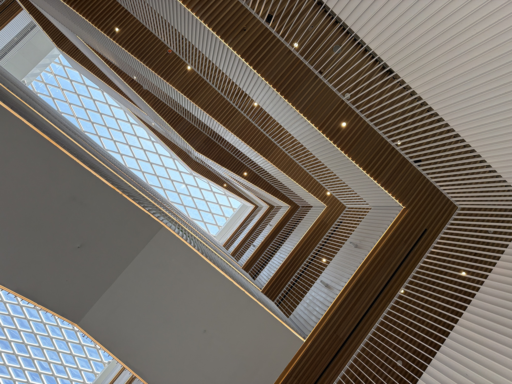 The photograph captures an interior view looking upwards from a multi-story atrium with a skylight. The atrium features floors with wooden paneling and white railings, illuminated by recessed lighting. The skylight is a diamond-shaped grid against a blue sky. The building appears modern and spacious. This architectural element is located at the Russian State Library in Moscow, Russia.