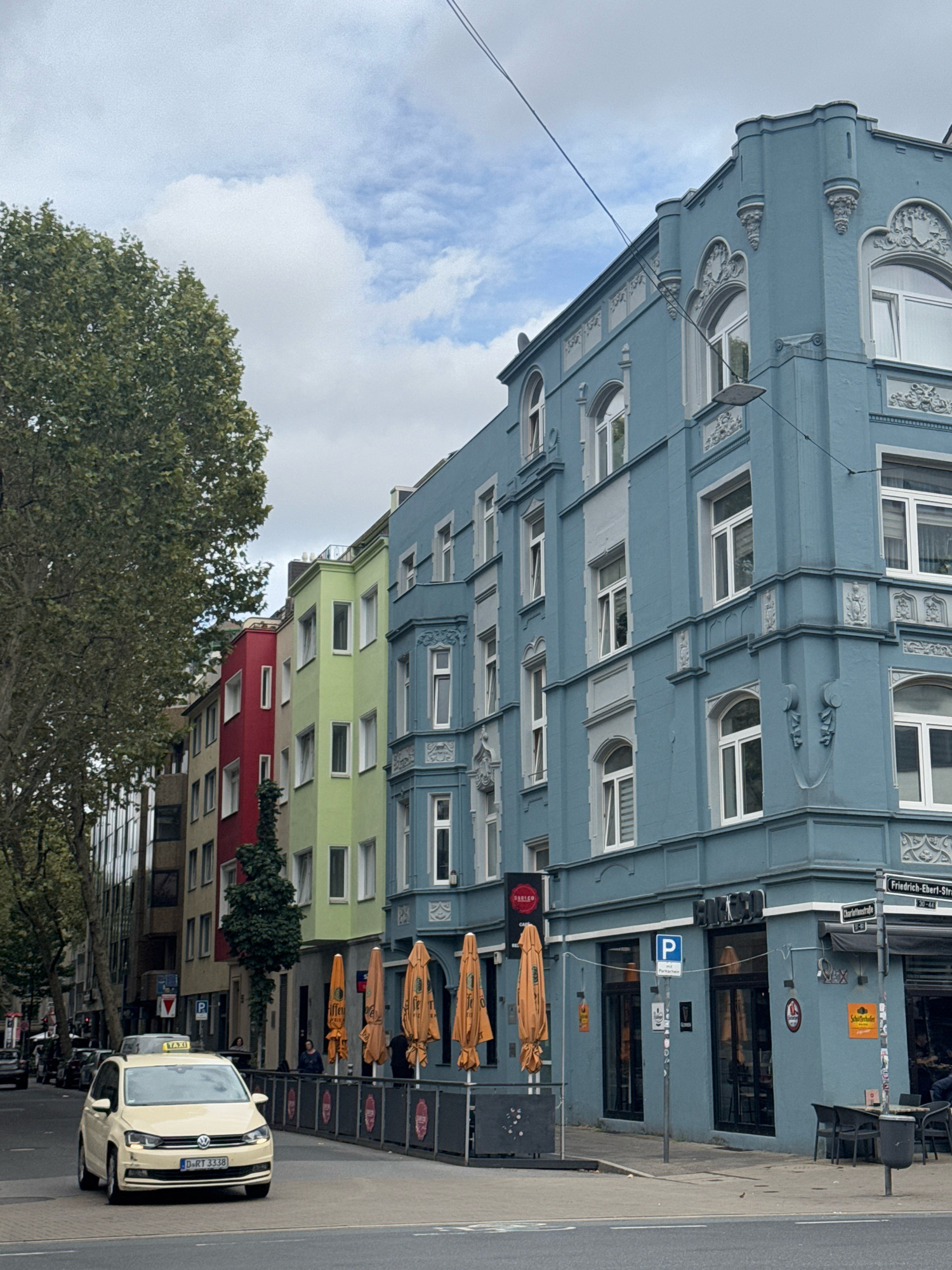 This is a street-level, medium-shot photograph taken in Dusseldorf, Germany, likely on a cloudy day. The image captures a row of colorful buildings lining a city street. The building on the right is a prominent, light blue structure with ornate architectural details, including arched windows and decorative molding. A street sign at the corner indicates "Friedrich-Ebert-Strasse" and "Charlottenstrasse."
In front of the blue building, there is an outdoor seating area with tables and chairs, shaded by closed orange umbrellas. A sign on the building reads "RAUSCO CAFE." Further down the street, a sequence of buildings are painted in vibrant colors: light green, red, and yellow, interspersed with neutral-toned buildings.
A yellow taxi is driving on the left side of the road, with its license plate visible as "D RT 3338." Several cars are parked or driving further down the street. A large tree with green foliage partially obstructs the view of the buildings on the left. People are visible sitting at tables outside the blue building and walking on the sidewalk. The sky is overcast with some patches of blue.