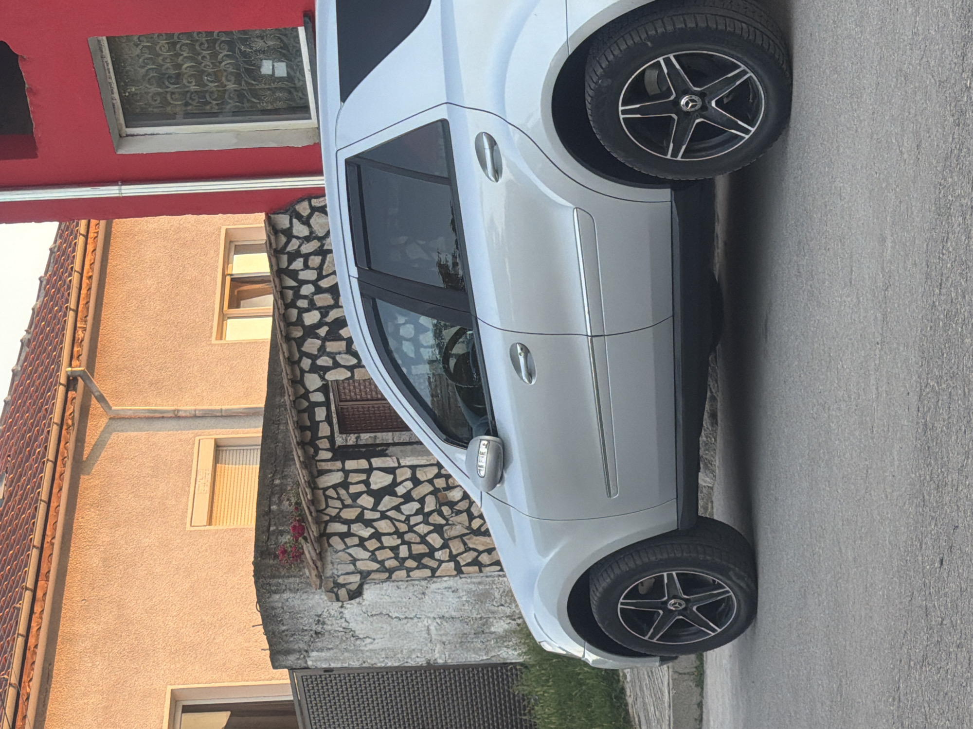 A silver Mercedes-Benz SUV is parked on a paved street in Pavel Banya, Bulgaria. The car is oriented vertically in the frame, with its wheels visible on the right side of the image. The vehicle has tinted windows and a metallic paint finish. It is parked in front of a building with a red facade and a dark, ornate window on the upper level. To the left of the red building is another building with a textured, light orange facade and a window with blinds. Part of a stone wall with irregular-shaped stones and some greenery is visible to the right of the orange building. The lighting suggests it is late afternoon or early evening, with the sun casting long shadows and creating warm tones on the buildings. The sky is not visible. There is no text visible in the image.