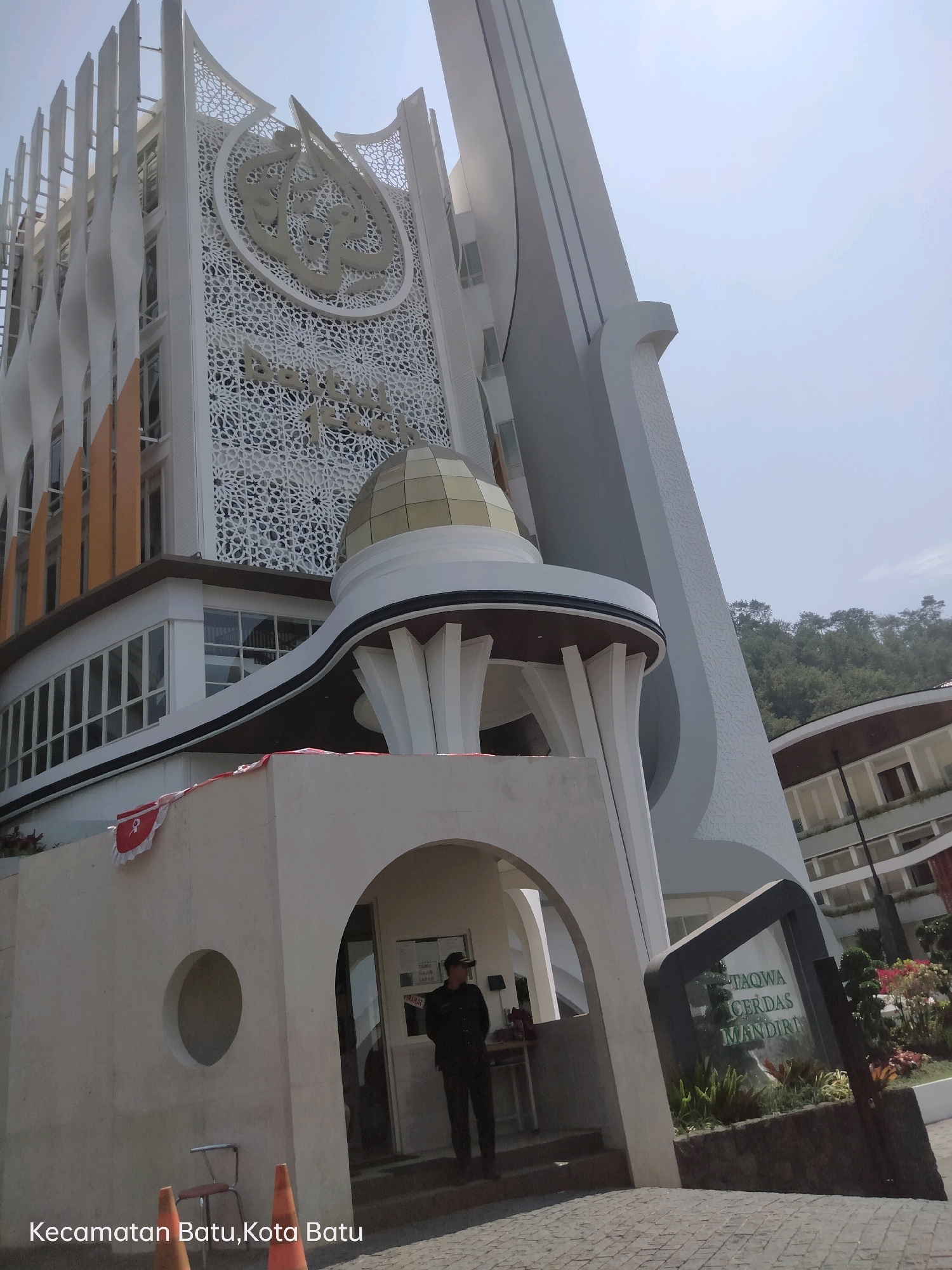 The image shows the front exterior of a modern building, likely a mosque or a religious educational institution, in the city of Batu, Indonesia. The building features a prominent white facade with intricate geometric patterns and Arabic calligraphy, a golden dome, and tall, curved architectural elements. In the foreground, a man in dark clothing and a cap stands by an entrance, possibly a security guard. Beside him, a sign with text in what appears to be Indonesian, "TAQWA CERDAS MANDIRI," is visible. Orange traffic cones and a chair are placed near the entrance. The scene is set against a backdrop of trees and hills, suggesting a location with natural surroundings. The text "Kecamatan Batu, Kota Batu" at the bottom indicates the administrative location.