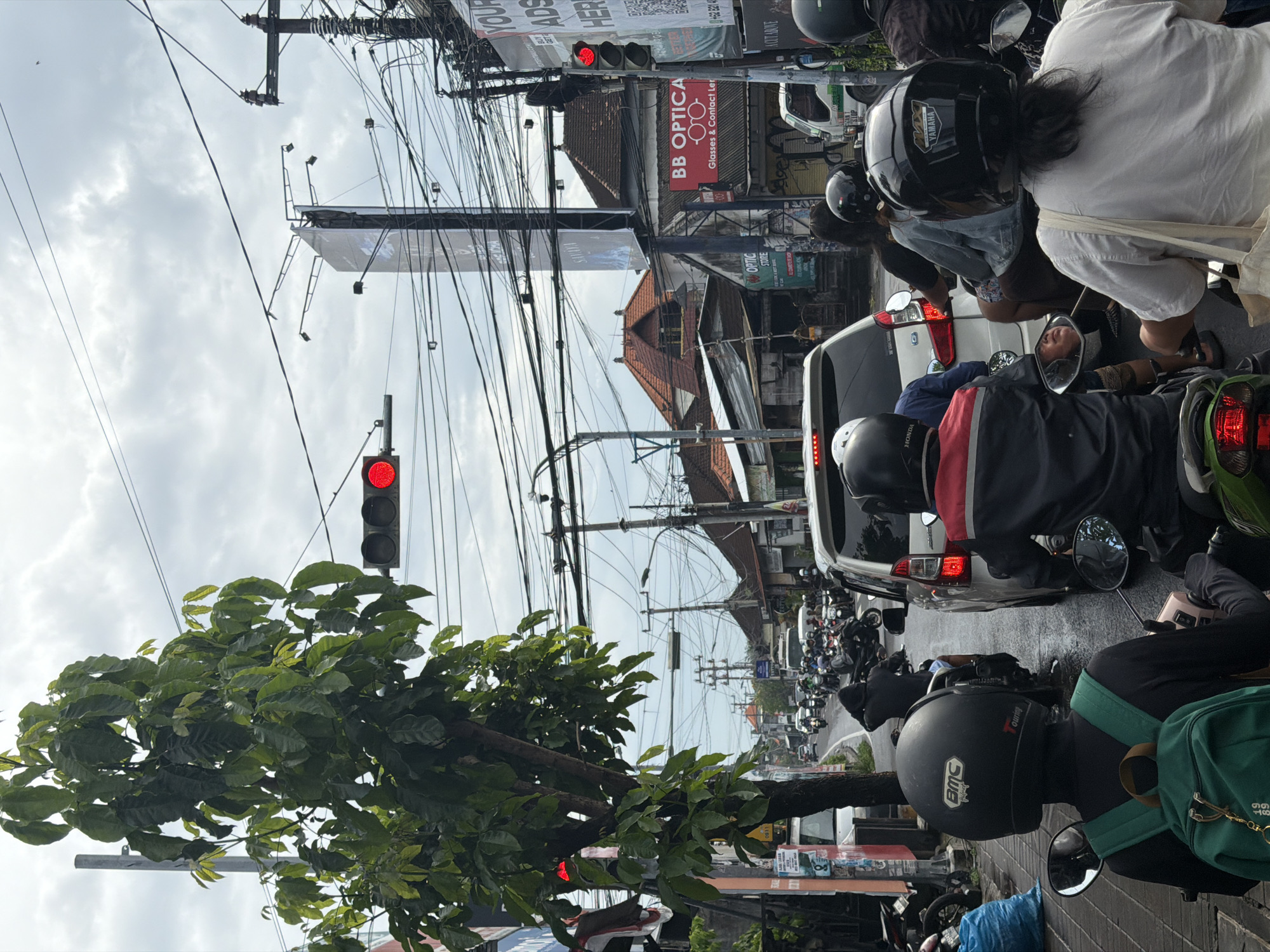 The image depicts a busy street scene in North Kuta, Indonesia, with traffic halted at a red light under an overcast sky. The perspective is from a low angle, looking forward along the street from behind a queue of vehicles.
Numerous motorcycles dominate the foreground and middle ground, with riders wearing helmets, waiting patiently for the traffic light to change. A white car is also part of the stopped traffic. In the immediate foreground, a large leafy tree partially obscures the lower section of a red traffic light. The sky is largely covered with clouds, suggesting an overcast day.
The background showcases a dense urban environment with various buildings, some featuring distinct traditional Indonesian architectural elements on their roofs. A complex network of overhead utility wires crisscrosses the sky, characteristic of many Southeast Asian cities. Billboards and various commercial signs are visible along the street.
Visible text includes:
* On shop signs: "BB OPTICA", "Glasses & Contact Ler" (likely Lenses), and "OPTIC STORE".
* On helmets and vehicles: "YAMAHA", "HONDA", "BMC", and "touring".
* On advertisements: "YOUR ADS HERE", "+62 8214" (part of a phone number), and "ACUT ABOVE".
* Other incidental text: "Seger", "0311", "18", and "99".
The scene captures a typical moment of urban congestion and daily life in a bustling Indonesian town.