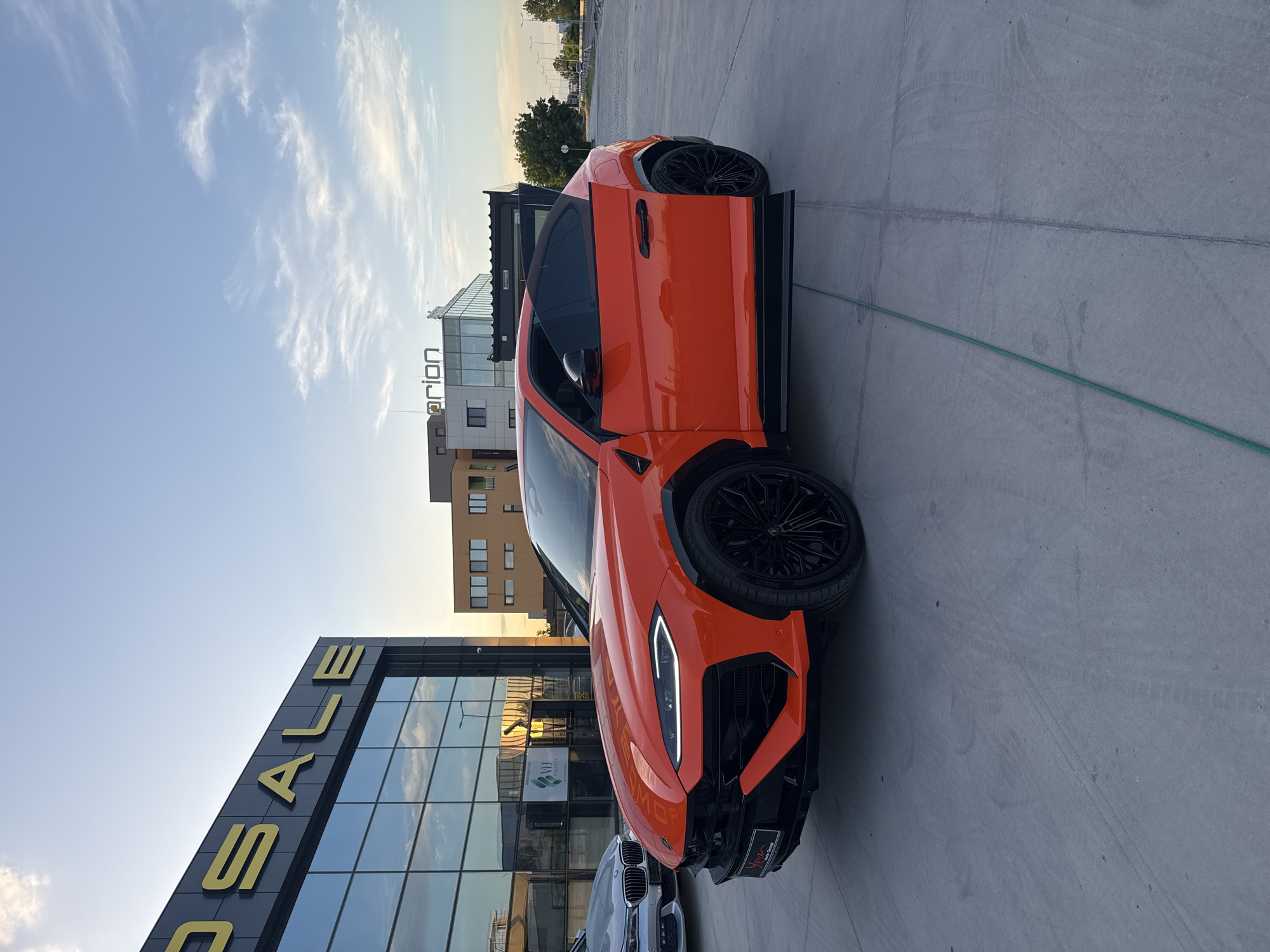 An orange Lamborghini Urus is parked outside a building with a large glass facade and the word "SALE" in yellow letters. In the background are buildings with architectural styles typical of Plovdiv, Bulgaria. The sky is partly cloudy. A silver-colored car is partially visible behind the Urus. There are no people visible in the frame. A thin green hose lies on the ground. The location is likely a car dealership or showroom.