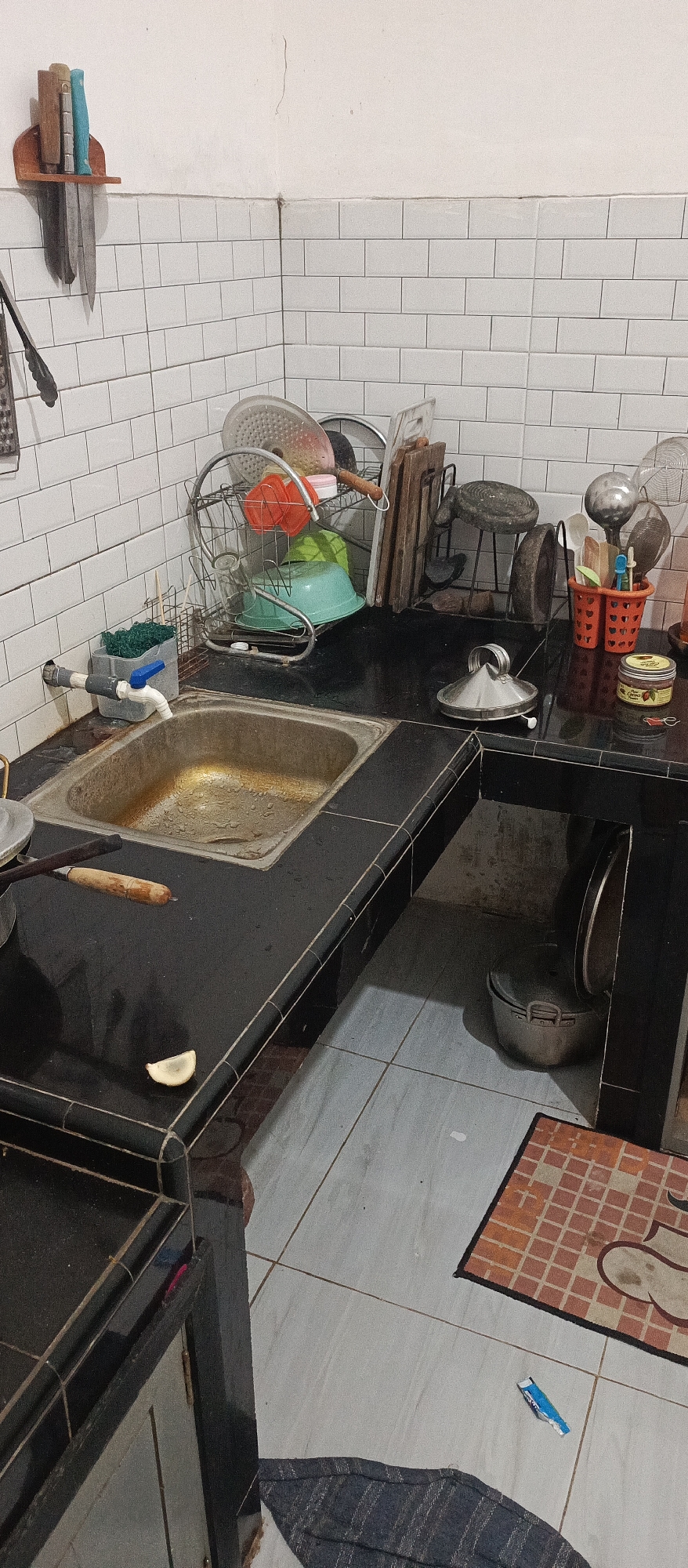 The image depicts a cluttered kitchen counter in what appears to be a residential setting in Arjasa, Indonesia. The counter, made of dark material with a tiled edge, is wet and shows signs of recent use. A stainless steel sink is partially visible, also appearing damp. On the counter, various kitchen items are present, including a drying rack with dishes, cutting boards, utensils in a red holder, and a small jar of what looks like jam. A wooden-handled knife is placed on the counter. In the lower right of the frame, a pot with a lid is stored under an overhang of the counter. The walls are tiled with white, rectangular tiles. The floor is tiled with light-colored squares and a patterned mat is visible near the bottom right corner. A blue wrapper is on the floor near the mat.