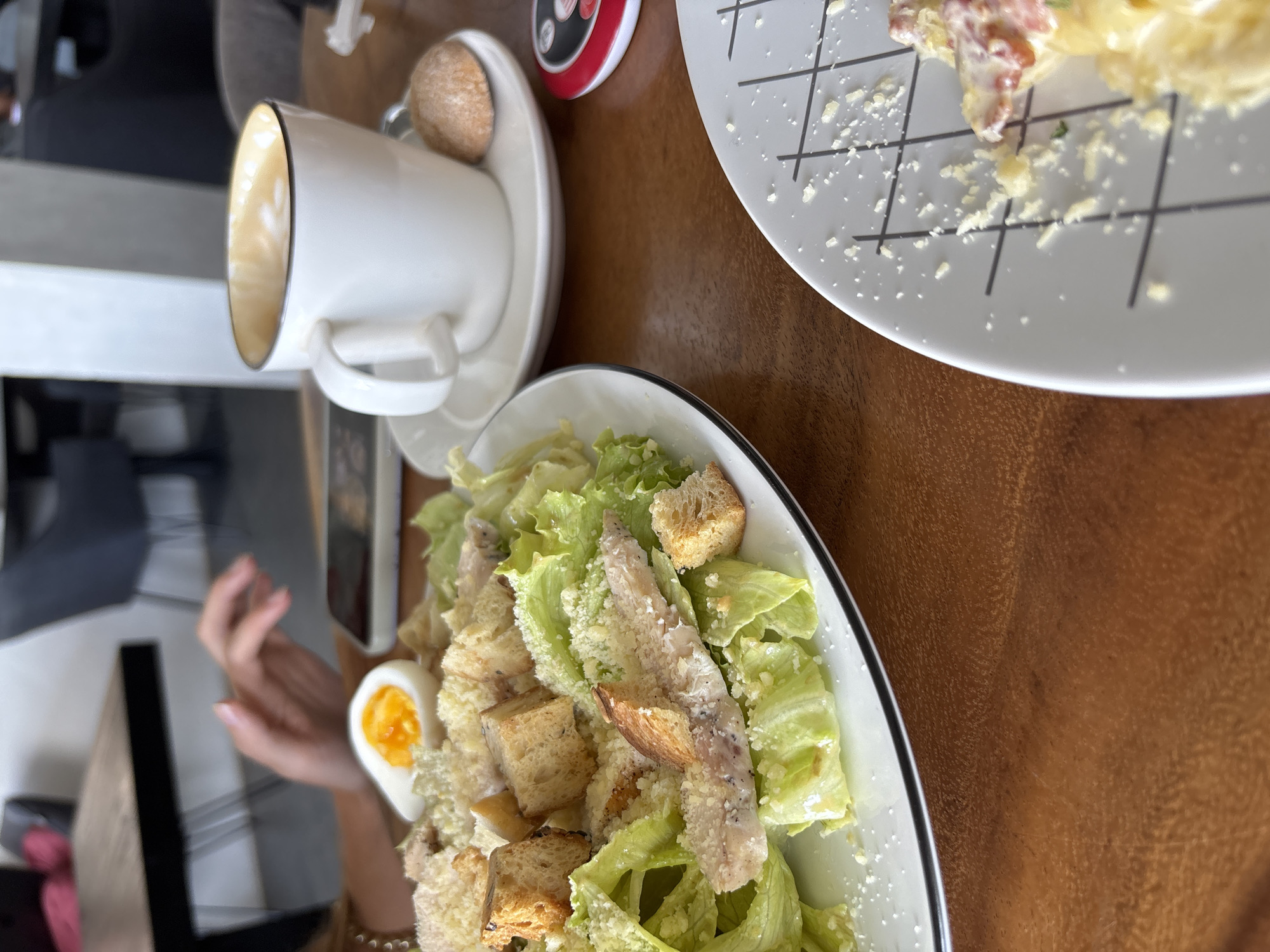 The image depicts a meal at a table in what appears to be a restaurant. There is a cup of coffee, two plates with food (likely salads, one possibly Caesar), a cell phone, and a woman's hand visible. The wooden tabletop suggests an indoor setting, while the presence of both a coffee cup and salads points towards a casual dining experience. Based on the specified location of Kuta Selatan, Indonesia, the setting is likely a cafe or restaurant catering to tourists or locals. The focus of the image is the food and drink on the table, indicating the subject is likely a food photograph taken during a meal.