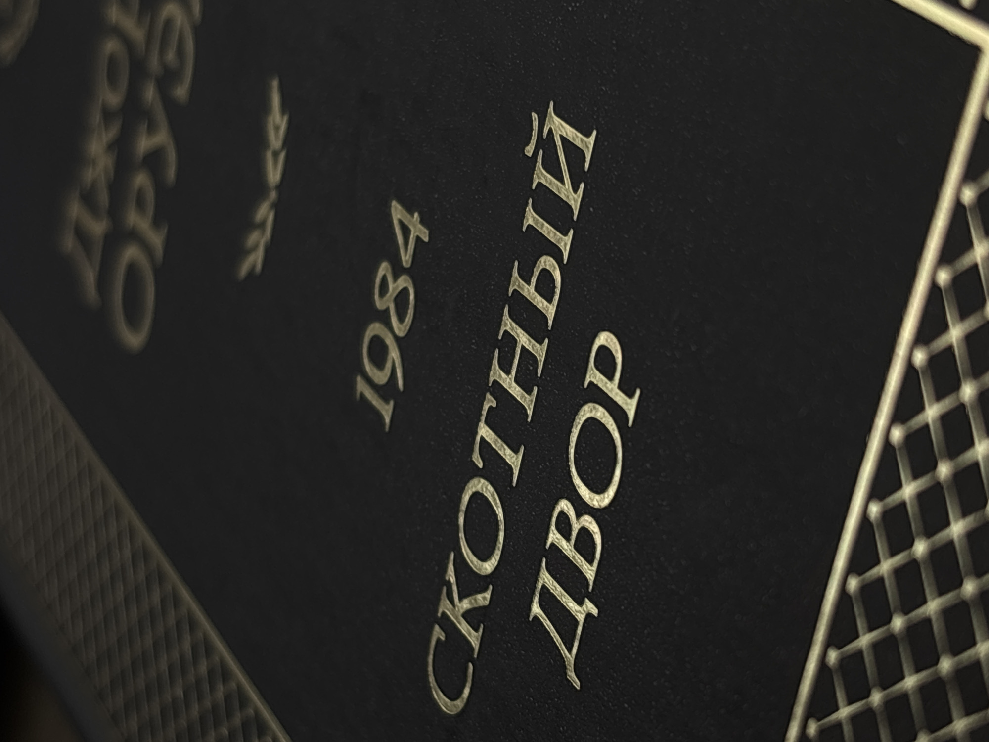 The image shows a close-up of a book cover with embossed gold lettering on a black textured background. The top of the cover features the name "ДЖОРДЖ ОРУЭЛЛ" (George Orwell) and a small laurel wreath emblem. Below this, the year "1984" is prominently displayed. The main title of the book, "СКОТНЫЙ ДВОР" (Animal Farm), is written in two lines in large, stylized gold letters. The bottom of the cover has a textured golden border with a diamond pattern. There is no indication that this image is located in Saint Petersburg, Russia.