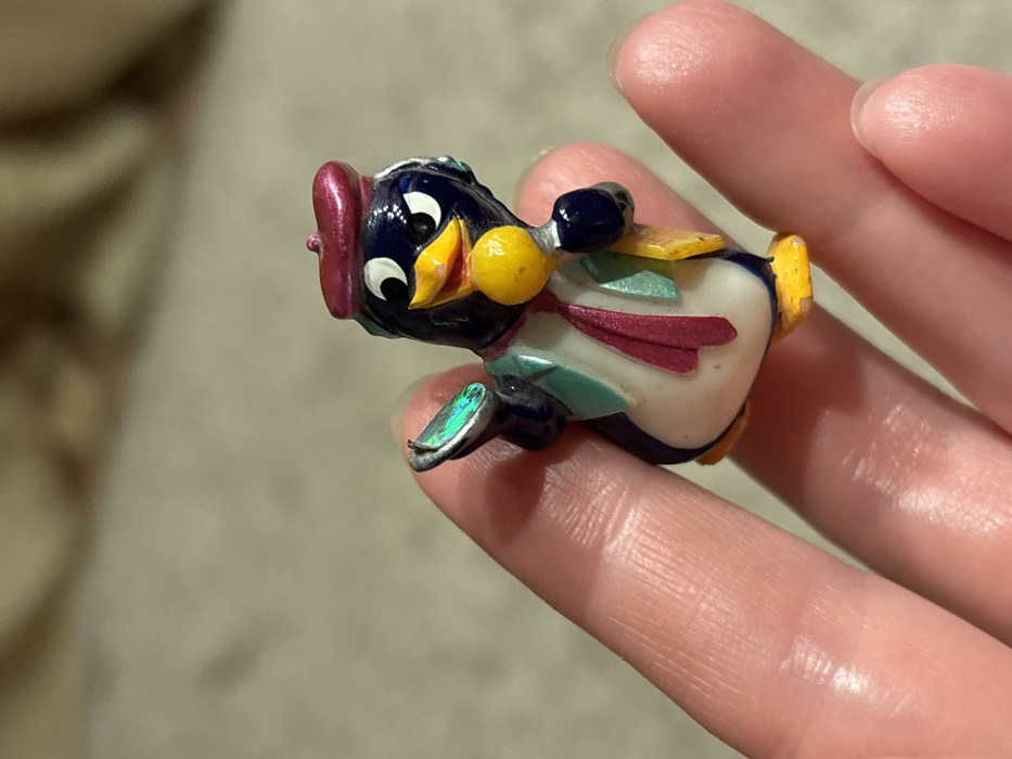 A colorful, decorative penguin figurine is held in a person's fingers. The penguin has a white belly, dark blue head and body, and a red hat. A yellow bow tie is attached to its neck. There are colored ribbons or accents in shades of teal and maroon on its chest. The penguin's beak is yellow, and one foot has a shimmery, iridescent finish. The background is blurry but appears to be a neutral color.