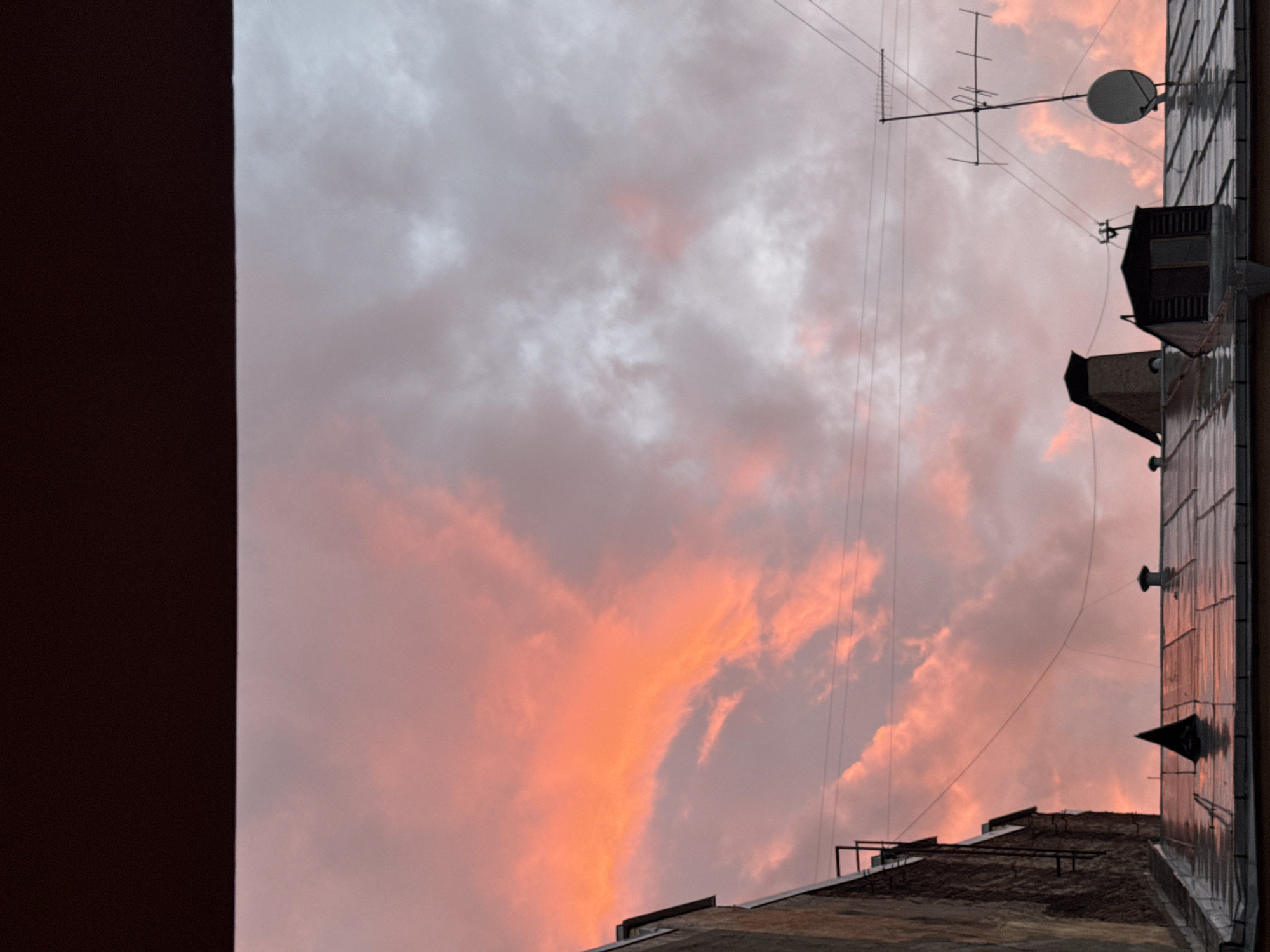 This is a medium-shot, horizontal photograph taken from a window looking out at a rooftop scene in Saint Petersburg, Russia. The sky is filled with dramatic, pink and orange clouds, suggesting either sunrise or sunset. In the foreground, the wet, metallic roof of a building is visible, with several dark, triangular chimney caps. To the left, the rough, brick wall of an adjacent building rises upwards, with a black metal fire escape attached. Further to the right, on the rooftop, are several smaller structures, including a pitched-roof building with a window and an older, brick chimney. An old-fashioned television antenna with a satellite dish is mounted on a pole, extending into the sky. The overall atmosphere is subdued, with the vibrant sky contrasting with the muted tones of the urban rooftop.