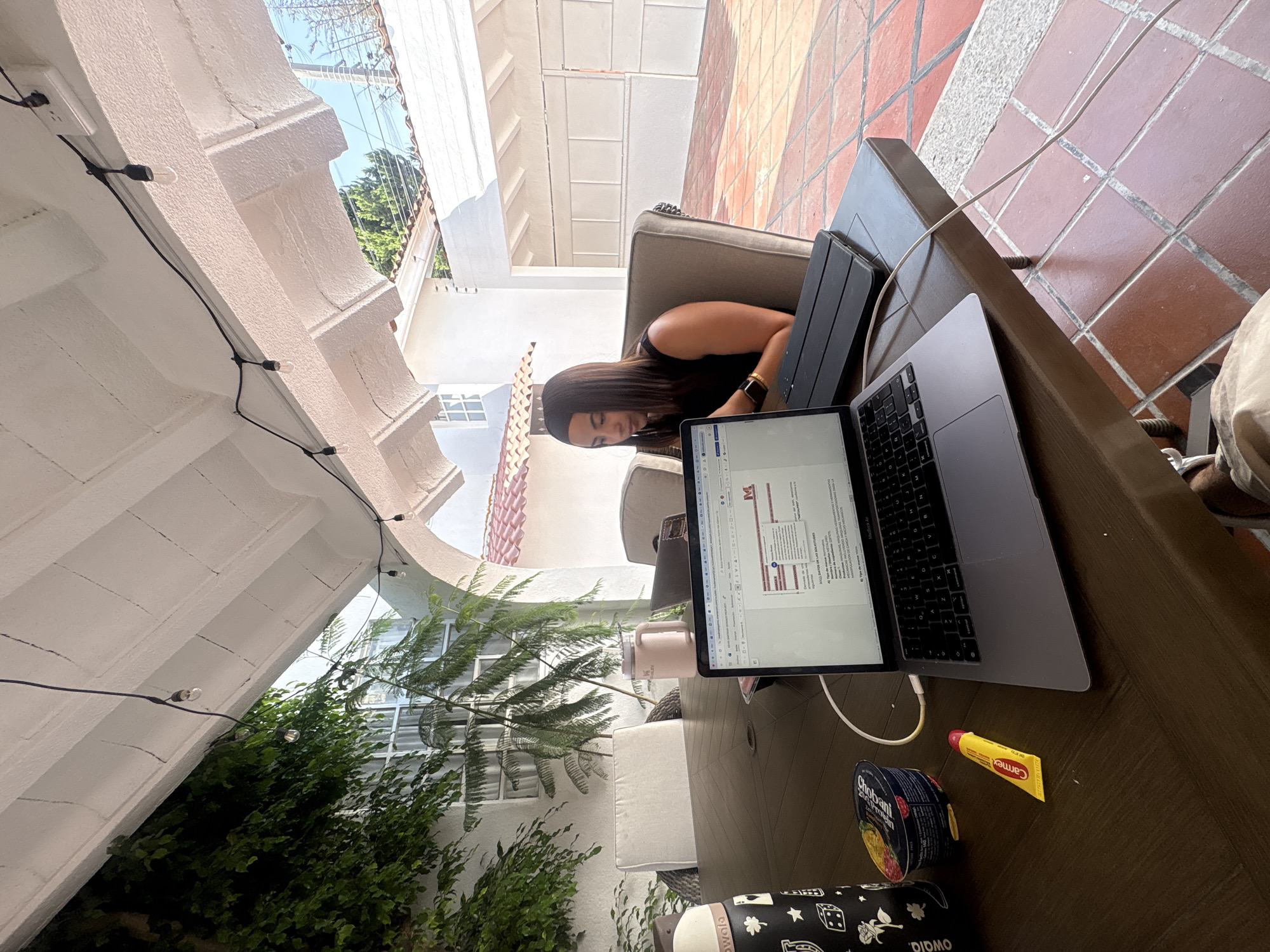 The photograph depicts a woman seated outdoors, working on a laptop computer. She has long, dark hair and is wearing a black tank top. She's positioned at a patio table where a laptop and a tablet are positioned, along with a container of Chobani yogurt, a tube of Carmex, and a Owala water bottle. The surroundings include patio furniture, tiled flooring, potted plants, and string lights. The architecture suggests a courtyard or patio space with a white archway and multiple windows. The image does not definitively confirm the location as Puebla, Mexico, but the architectural style is consistent with colonial Mexican architecture found in Puebla.